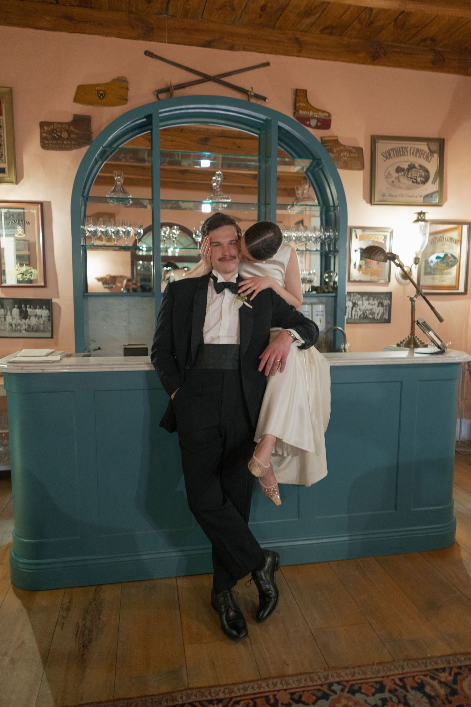 Groom in tuxedo leans against teal bar counter while bride in white dress embraces him in vintage decorated venue.
