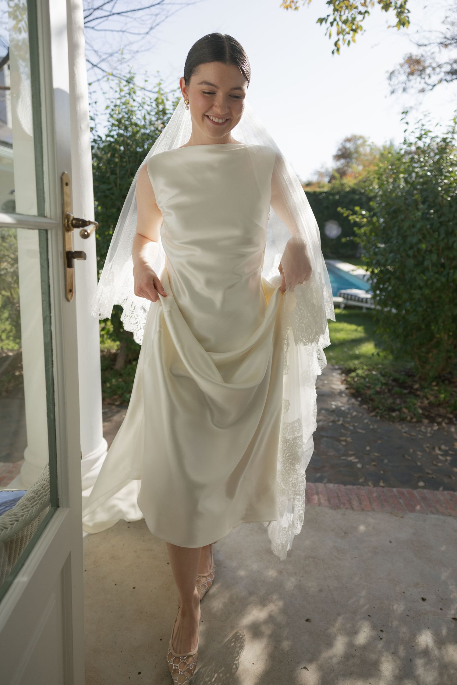 Bride in ivory satin wedding dress and lace veil lifts skirt while standing in sunlit doorway.