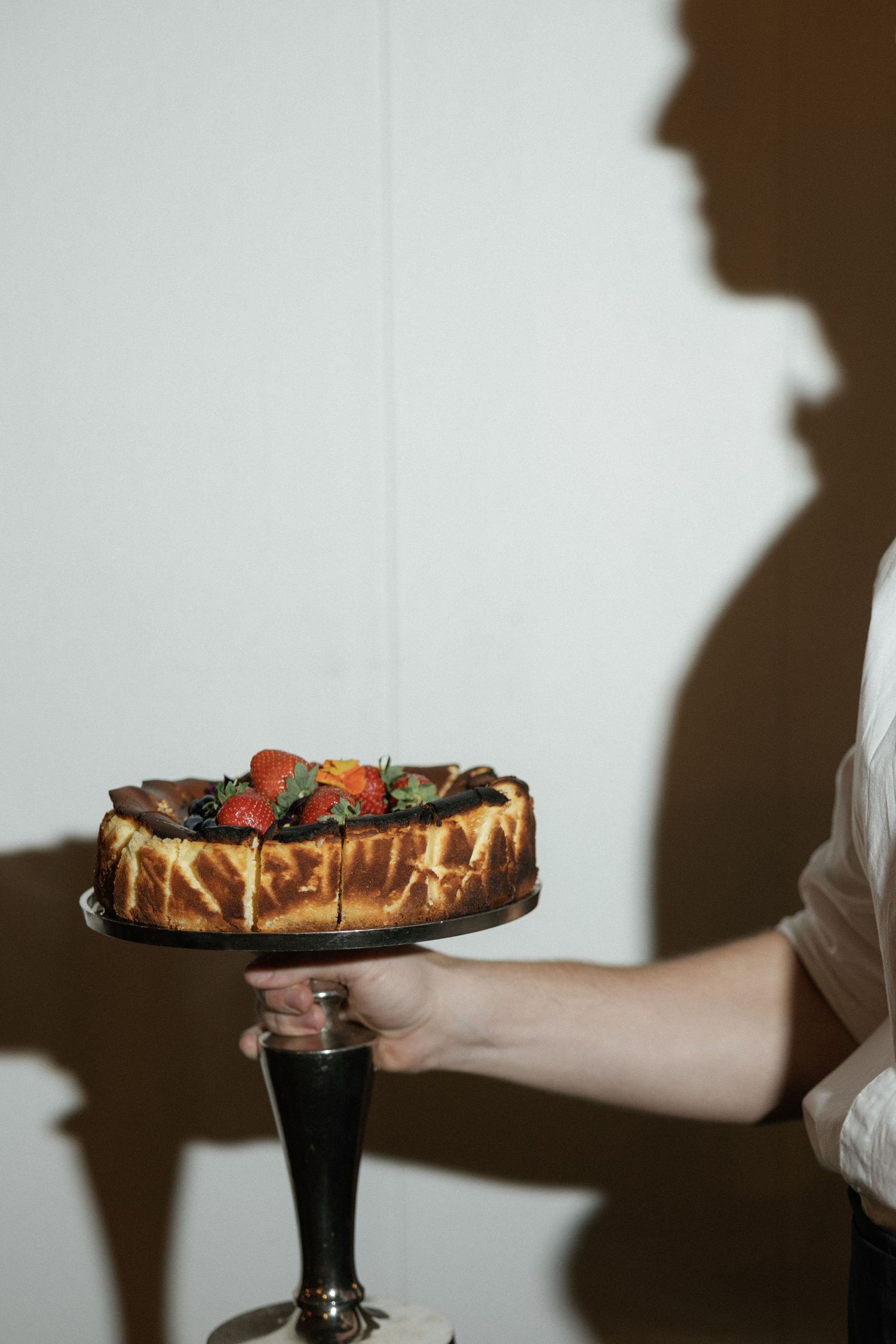 Person holding a cheesecake topped with fresh strawberries on an elegant cake stand, with a shadow silhouette on white wall.