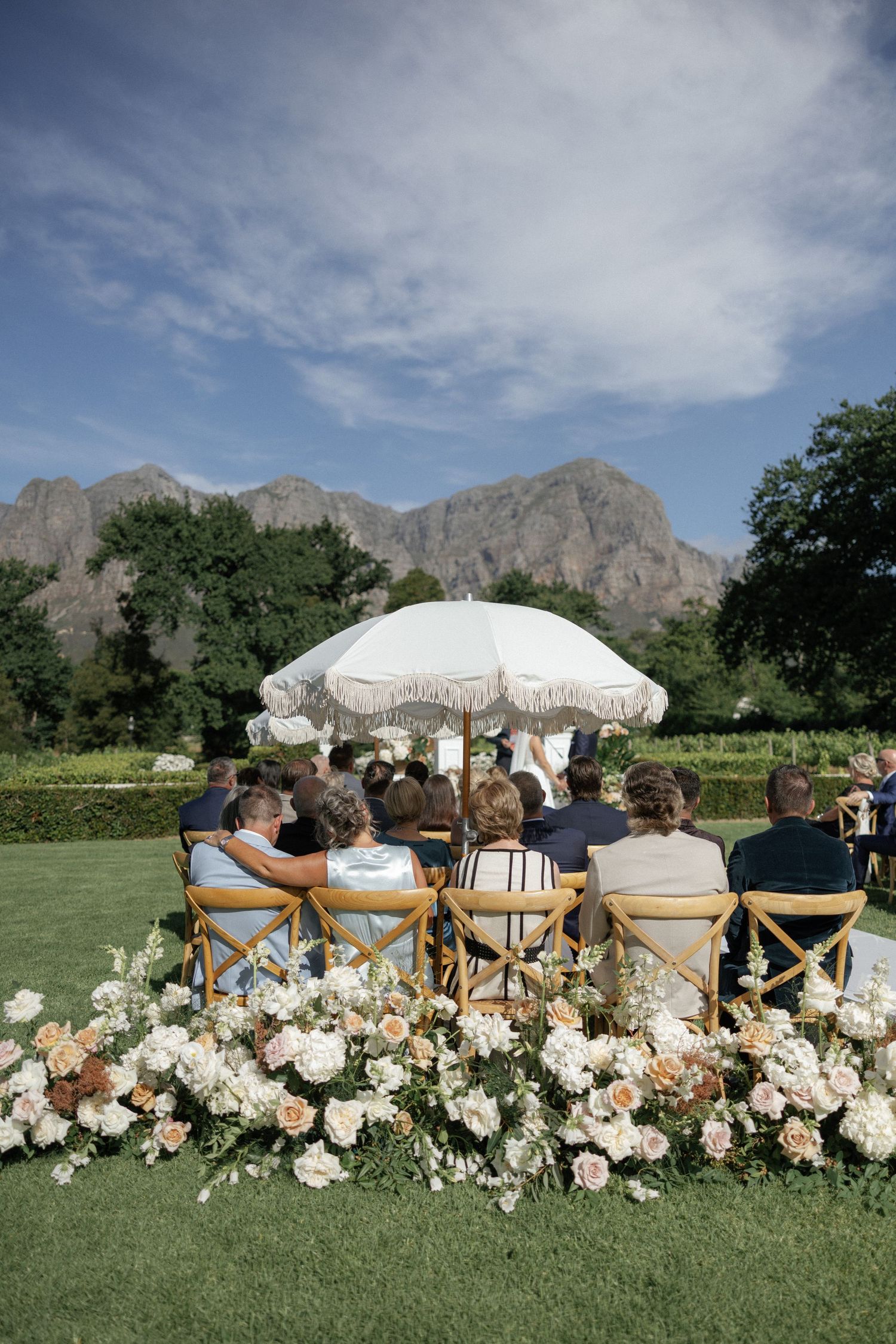 Outdoor wedding ceremony with guests seated on wooden chairs, white floral arrangements, fringed umbrella, and mountain backdrop.
