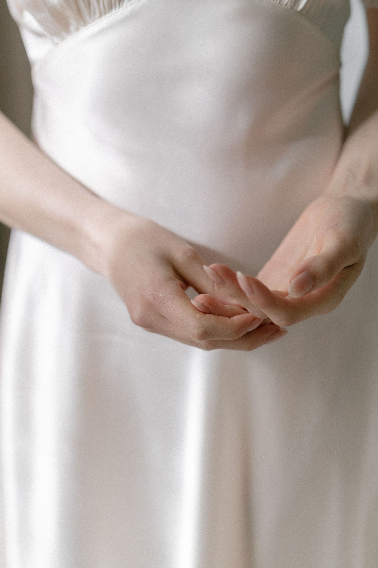 Close-up of bride in white satin wedding dress gently clasping her hands together.