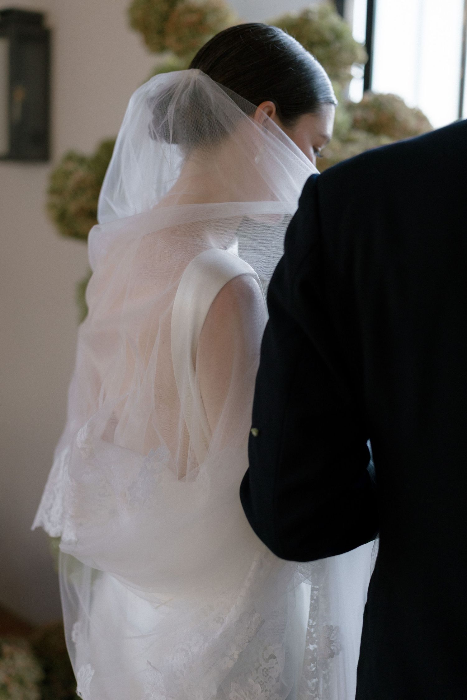 Close-up back view of bride in sleeveless white gown with flowing veil standing beside groom in chapel.