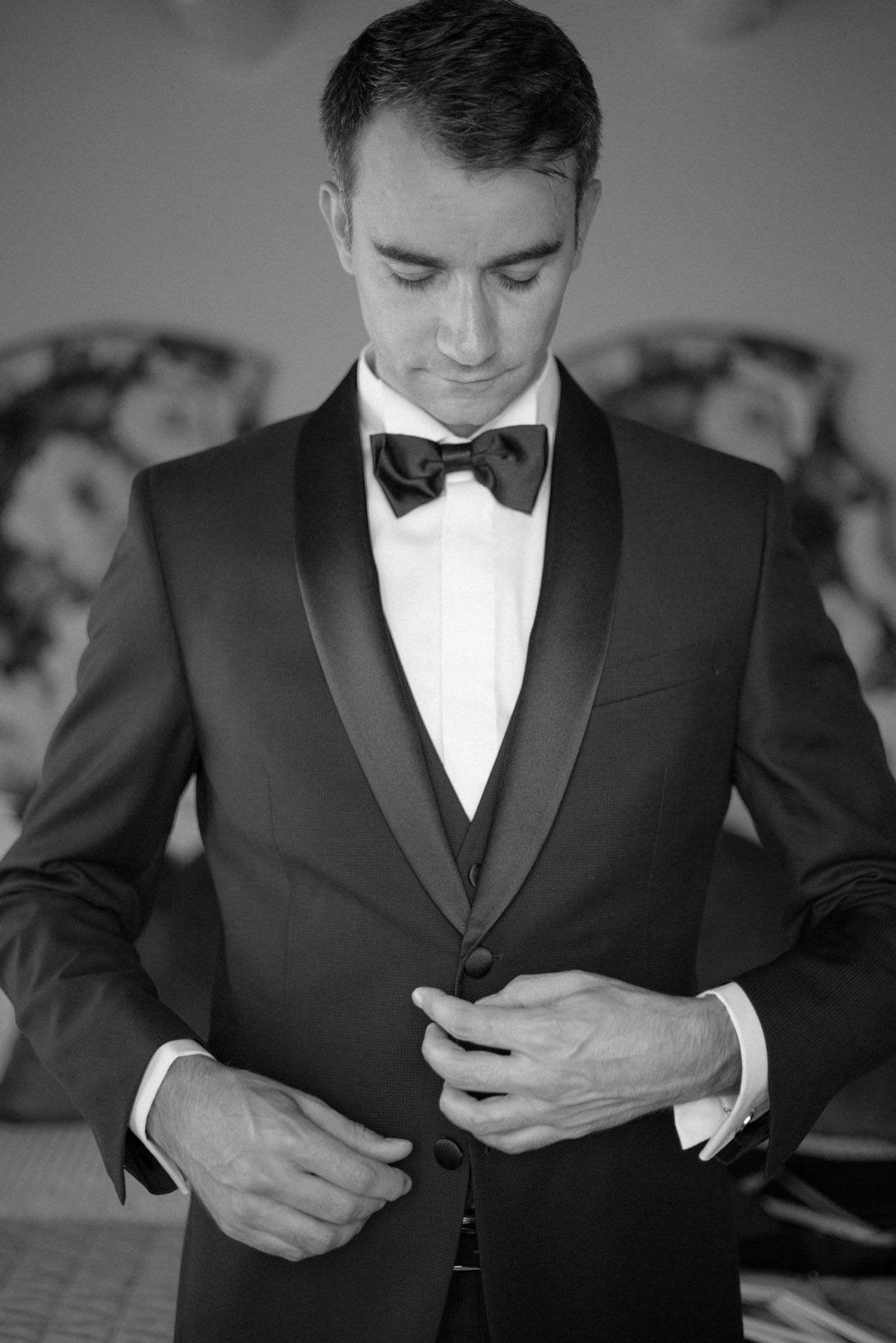 Black and white portrait of groom in tuxedo buttoning jacket before wedding ceremony.
