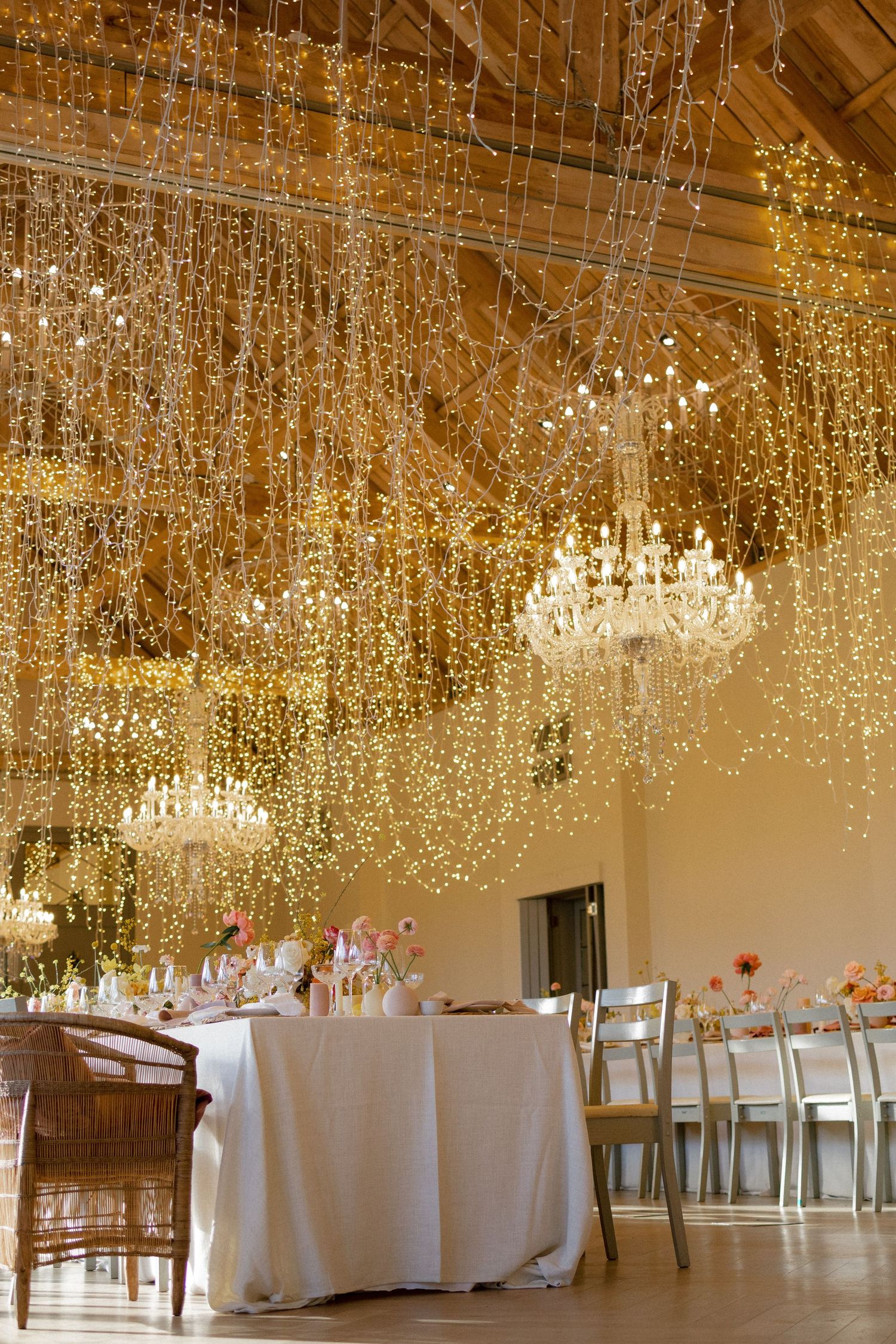 Elegant wedding reception hall with cascading fairy lights ceiling and crystal chandeliers above white draped tables.