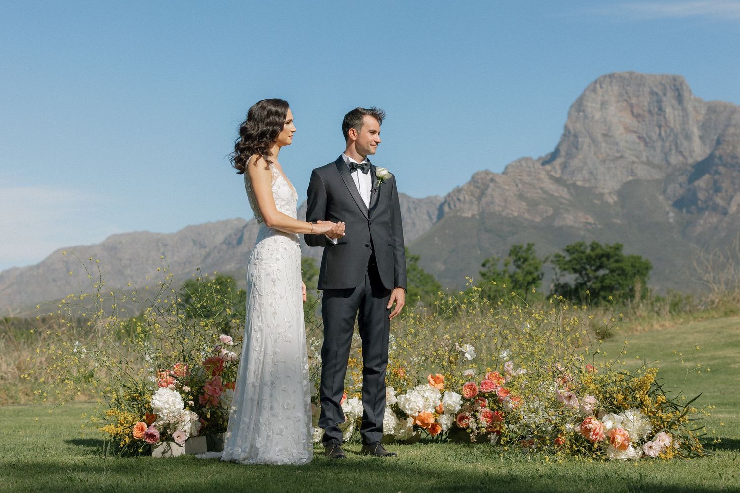 Bride and groom holding hands at outdoor ceremony altar with stunning mountain peak backdrop and wildflower fields.