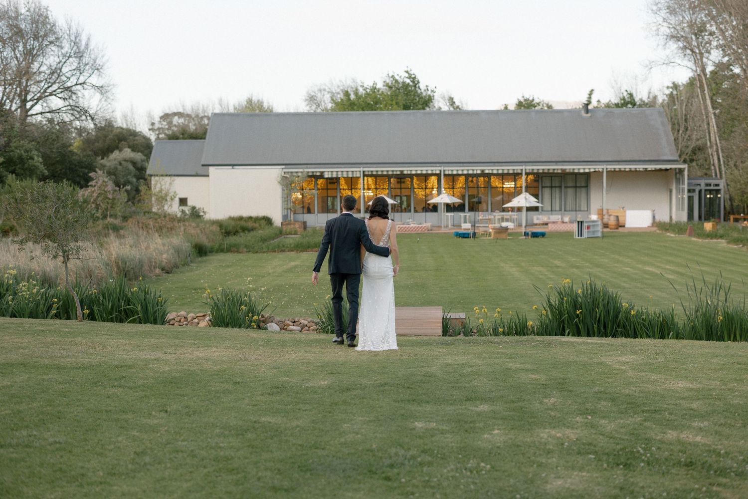 Bride and groom walking hand in hand toward illuminated wedding venue at dusk.