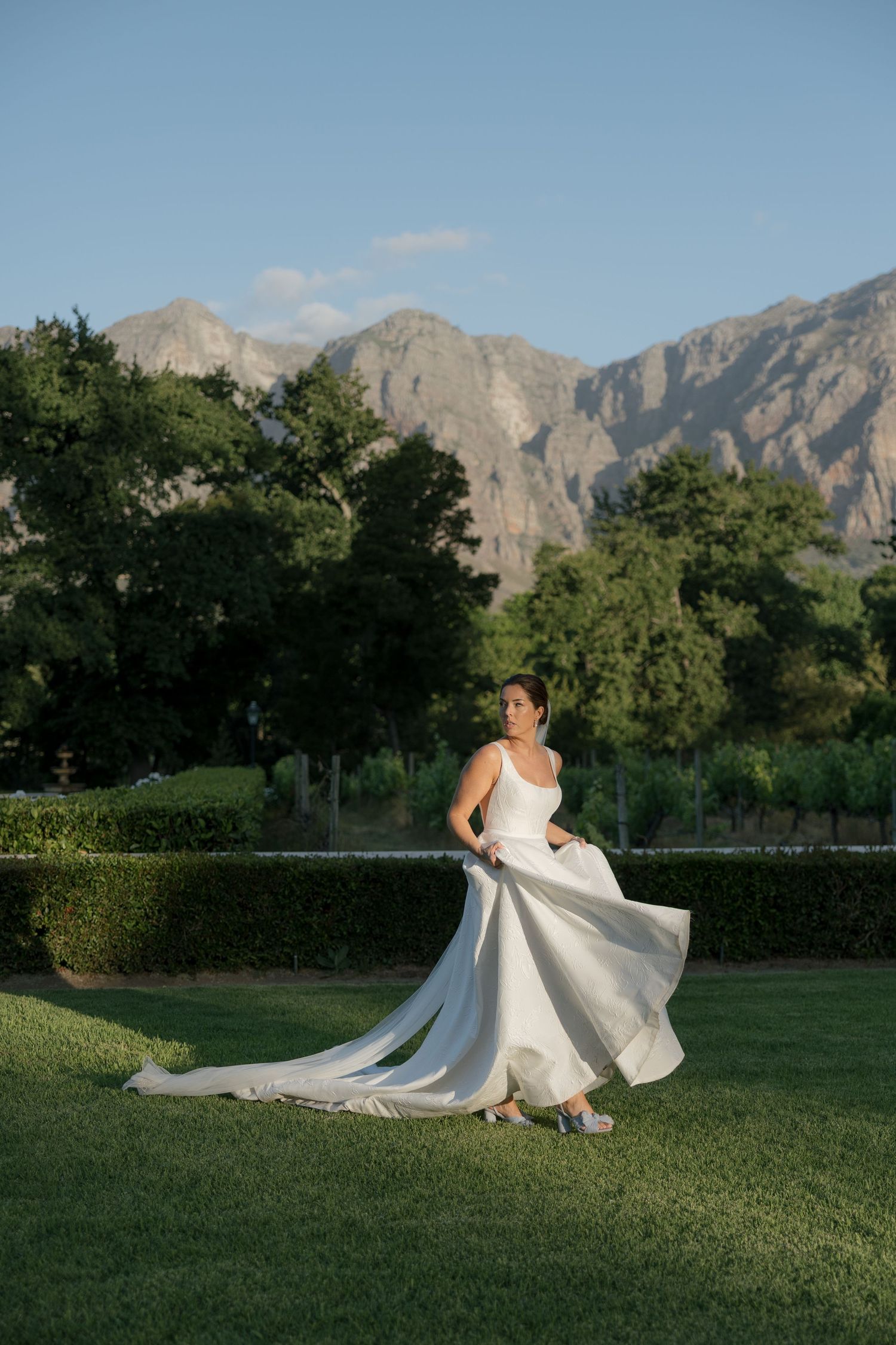 Bride in elegant white gown with long train twirling on green lawn with mountain backdrop at golden hour.