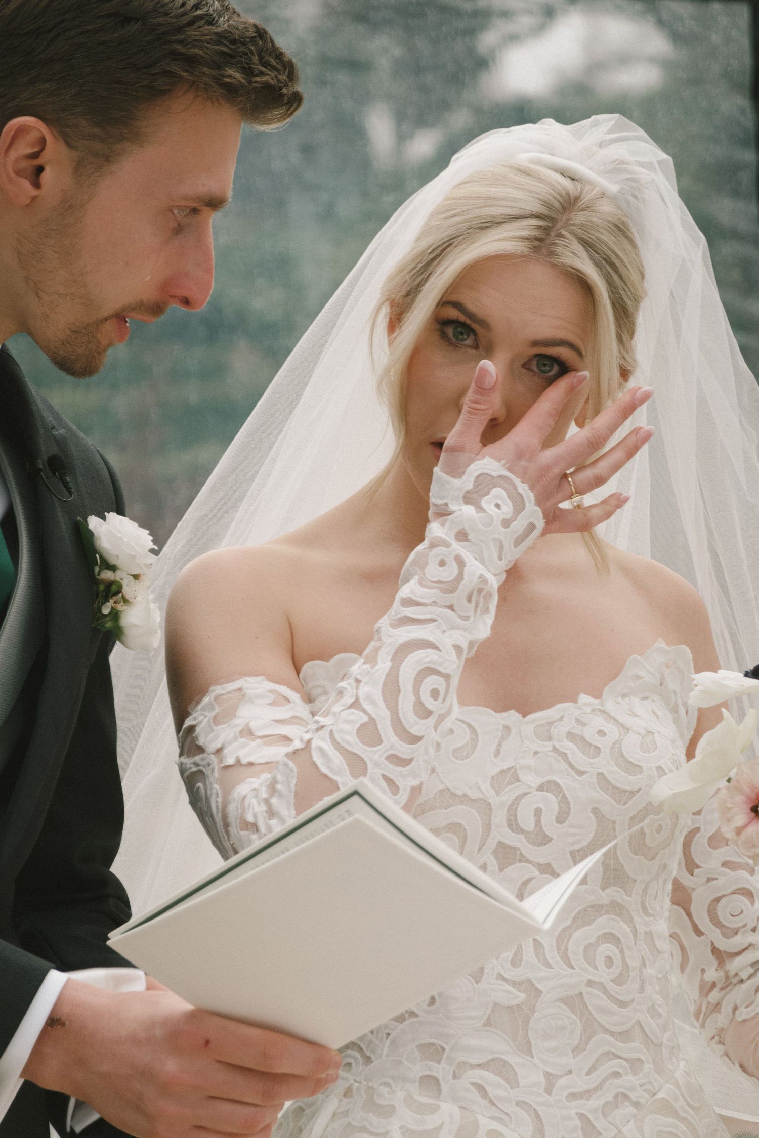 Emotional bride in lace gown and veil wipes tears while groom reads wedding vows during outdoor ceremony.