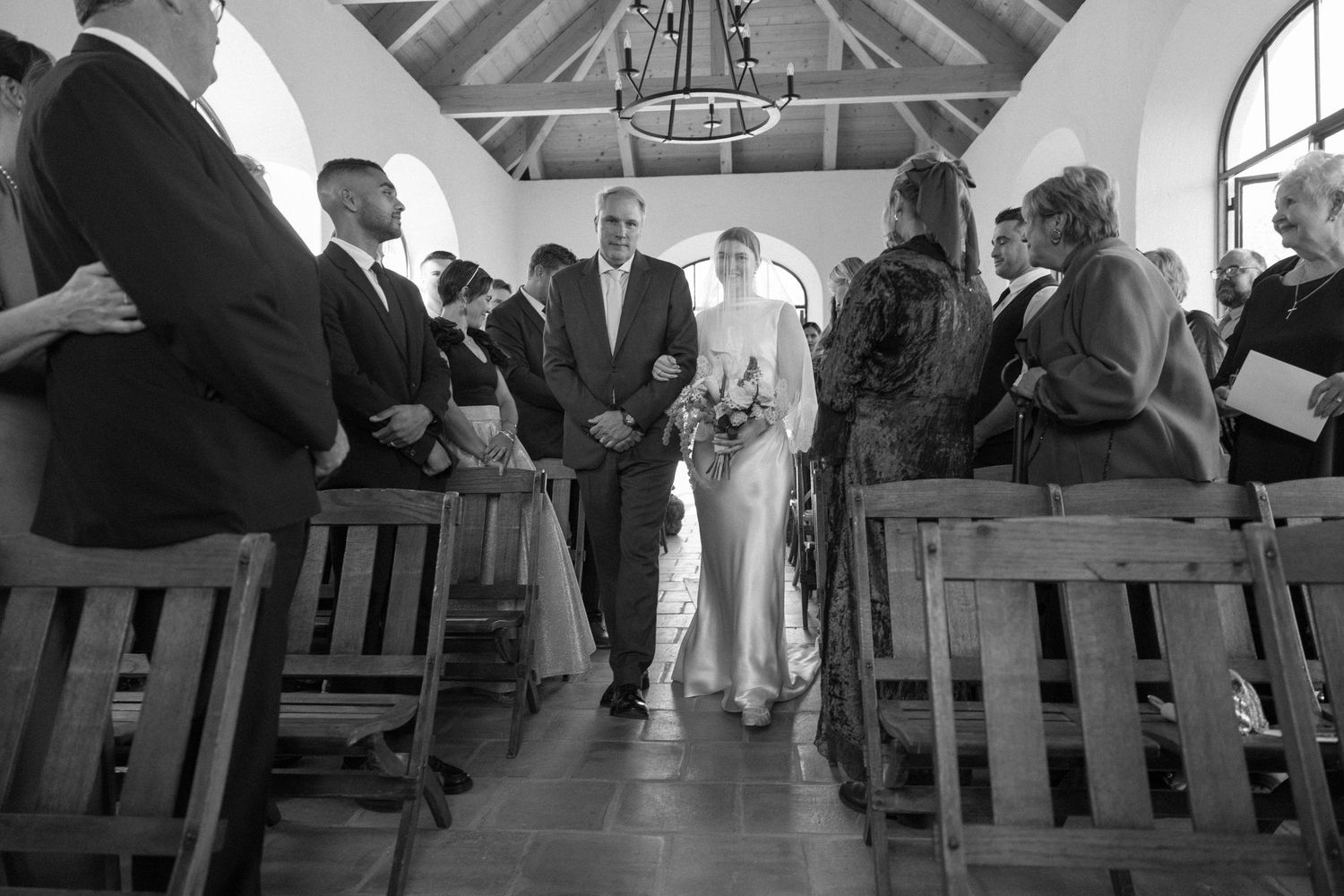 Black and white photo of bride walking down chapel aisle with escort past seated wedding guests.
