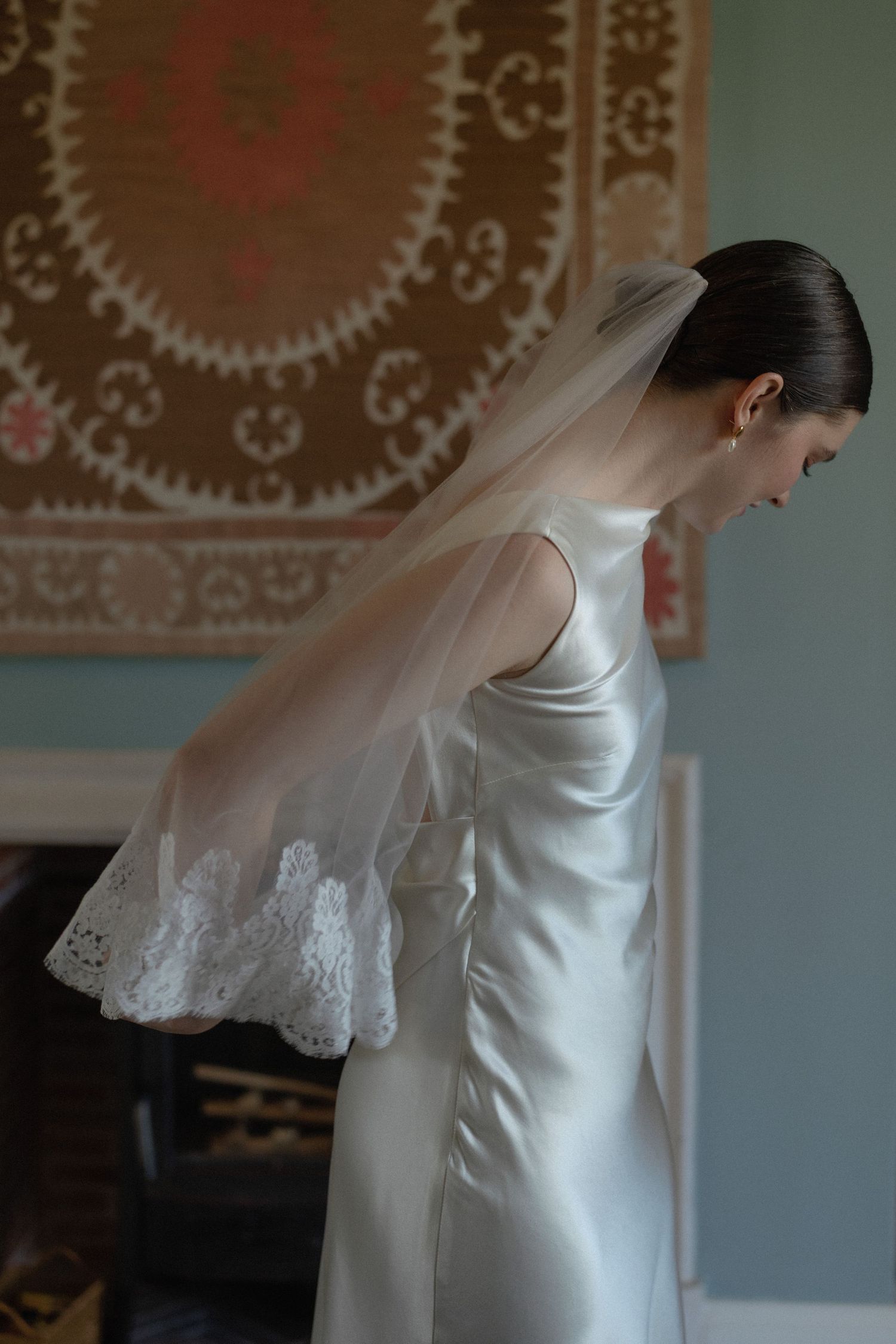 Bride in sleek white satin gown with lace-trimmed veil bows head in softly lit interior room.