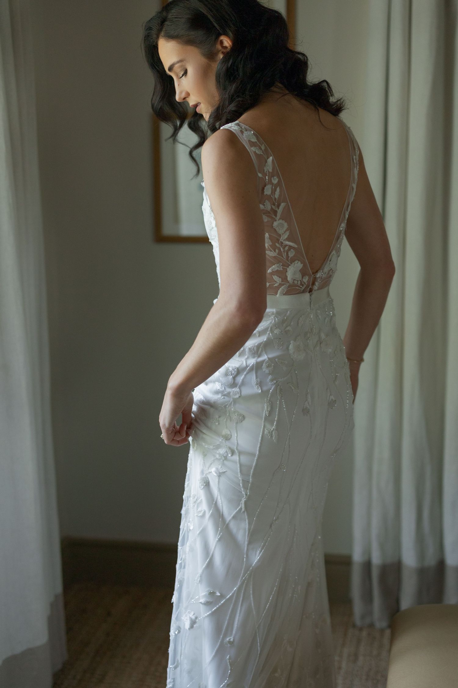 Bride in backless floral lace wedding gown posing elegantly in a softly lit hotel room.