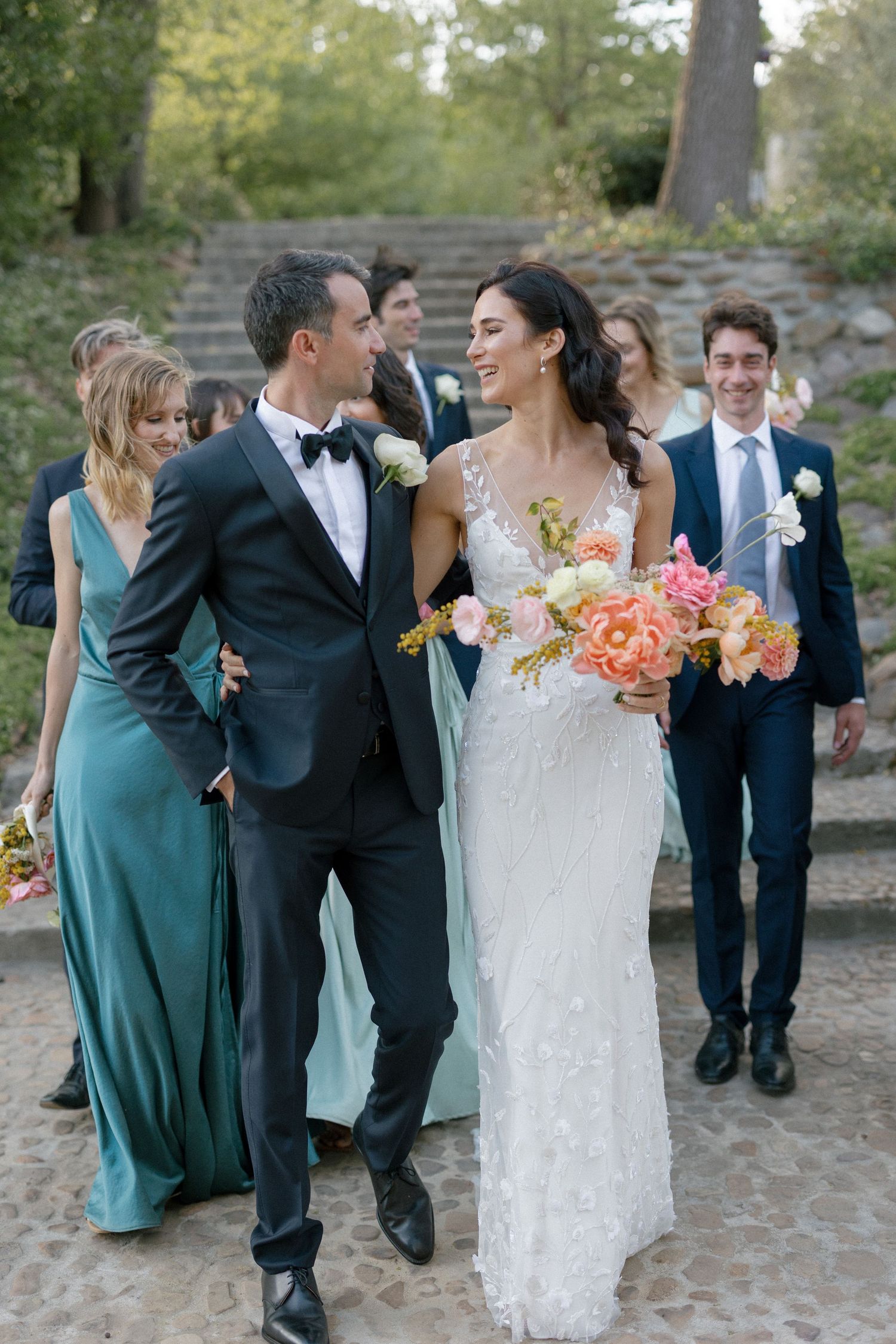 Bride and groom walking with wedding party, bride holding colorful peach and pink bouquet.