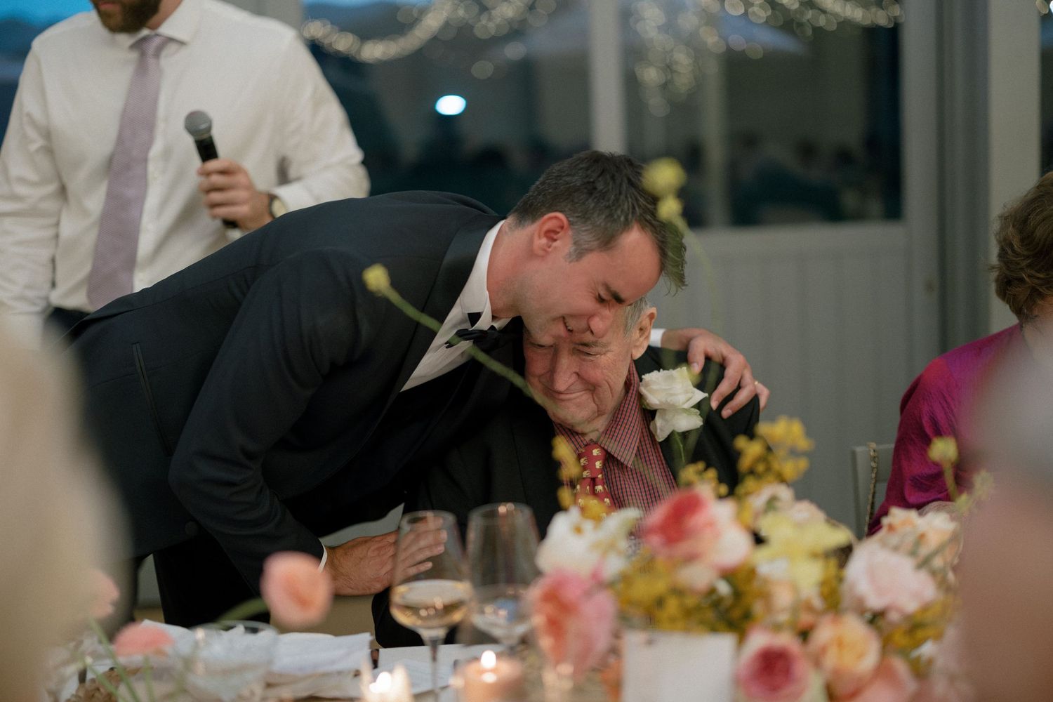 Two men share an emotional embrace at a wedding reception dinner table with floral centerpieces.