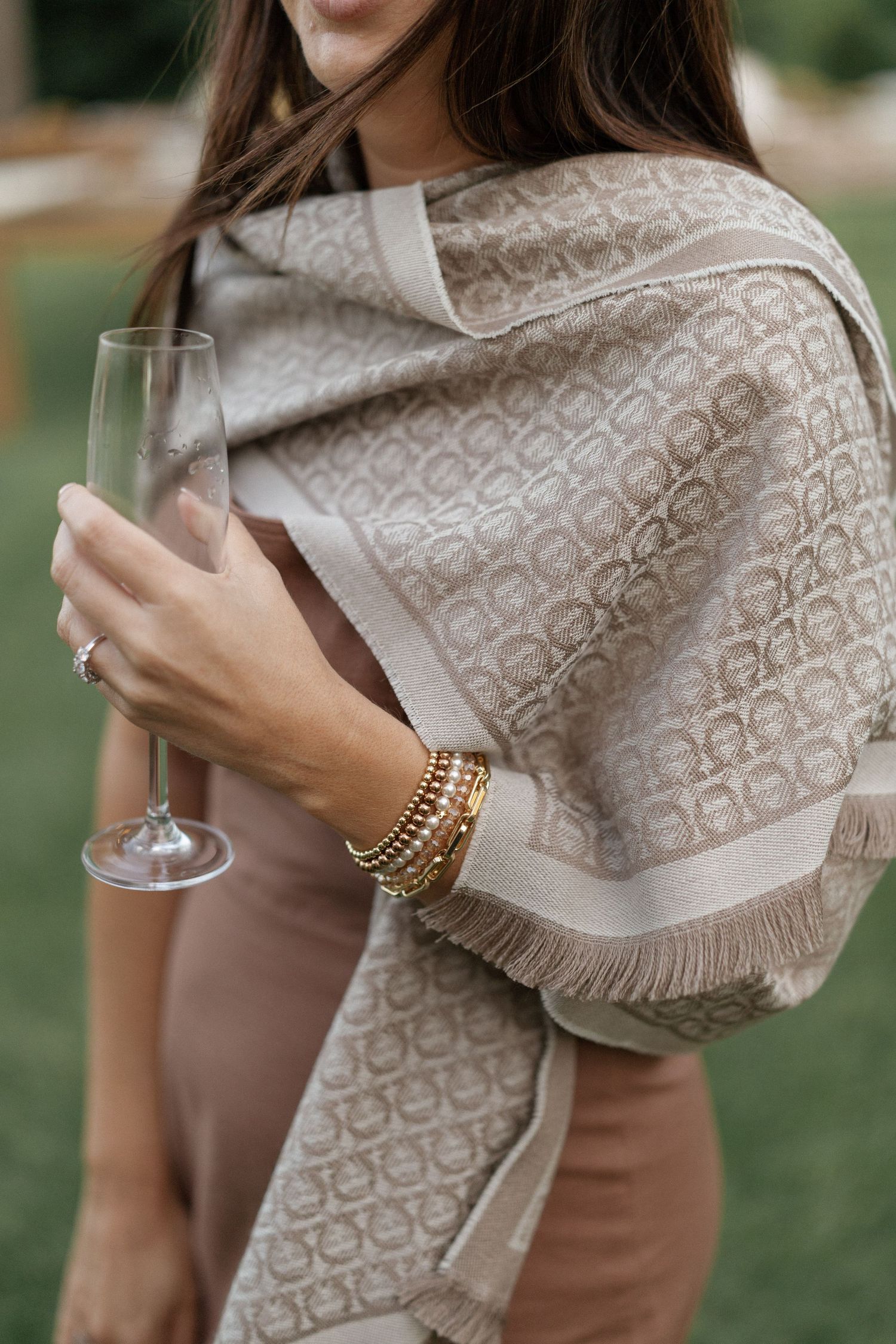 Woman in beige dress holding champagne flute, wearing gold bracelets and elegant taupe patterned fringe shawl outdoors.