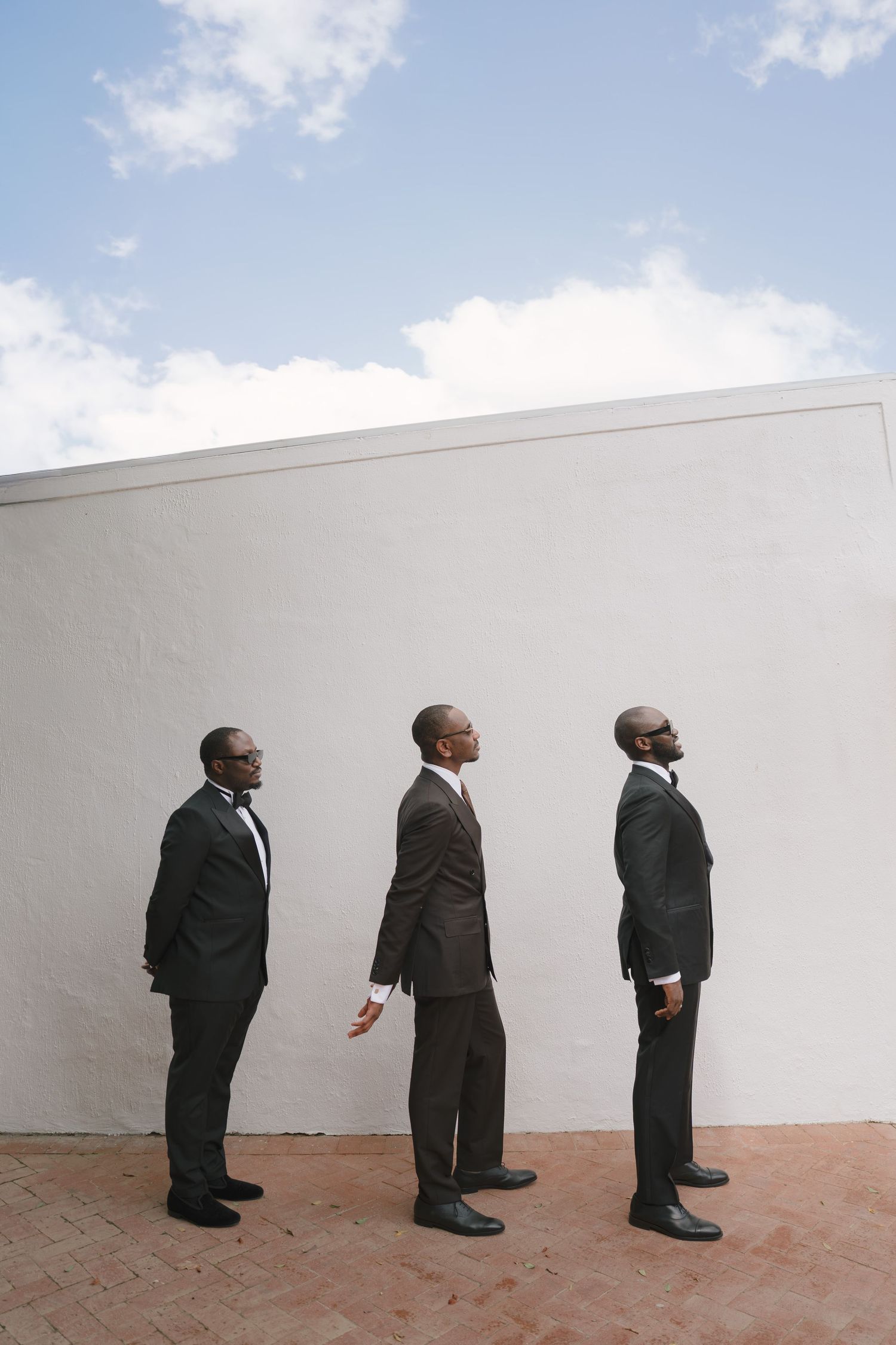 Three Black businessmen in dark suits standing in profile against a white wall, looking upward toward a cloudy sky.