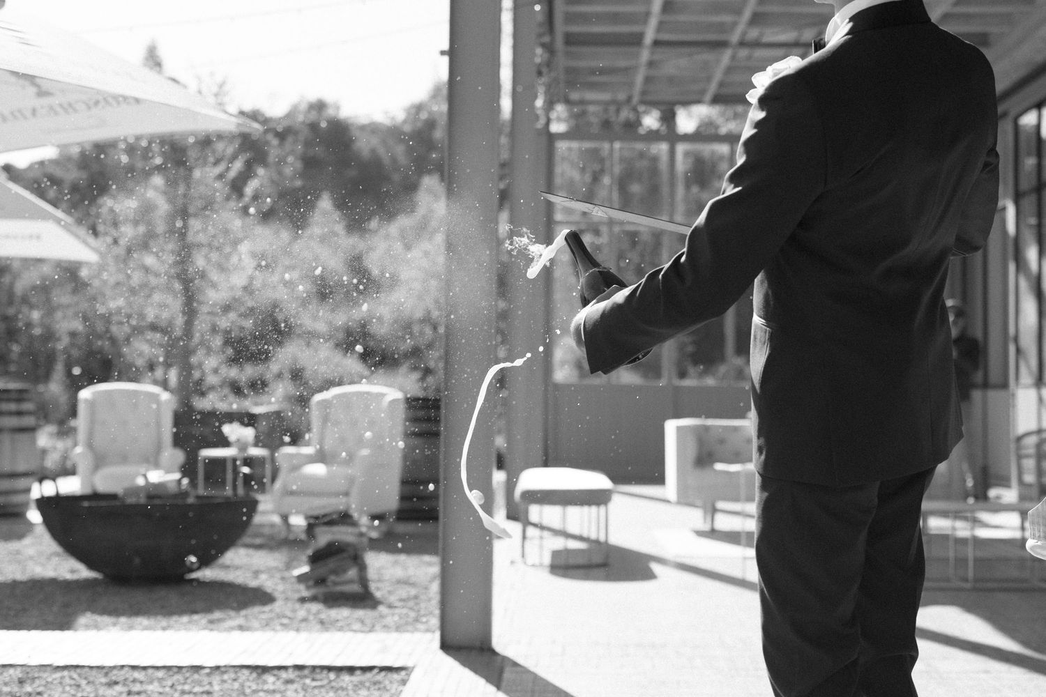 Black and white photo of person in suit sabering champagne bottle at outdoor wedding venue.