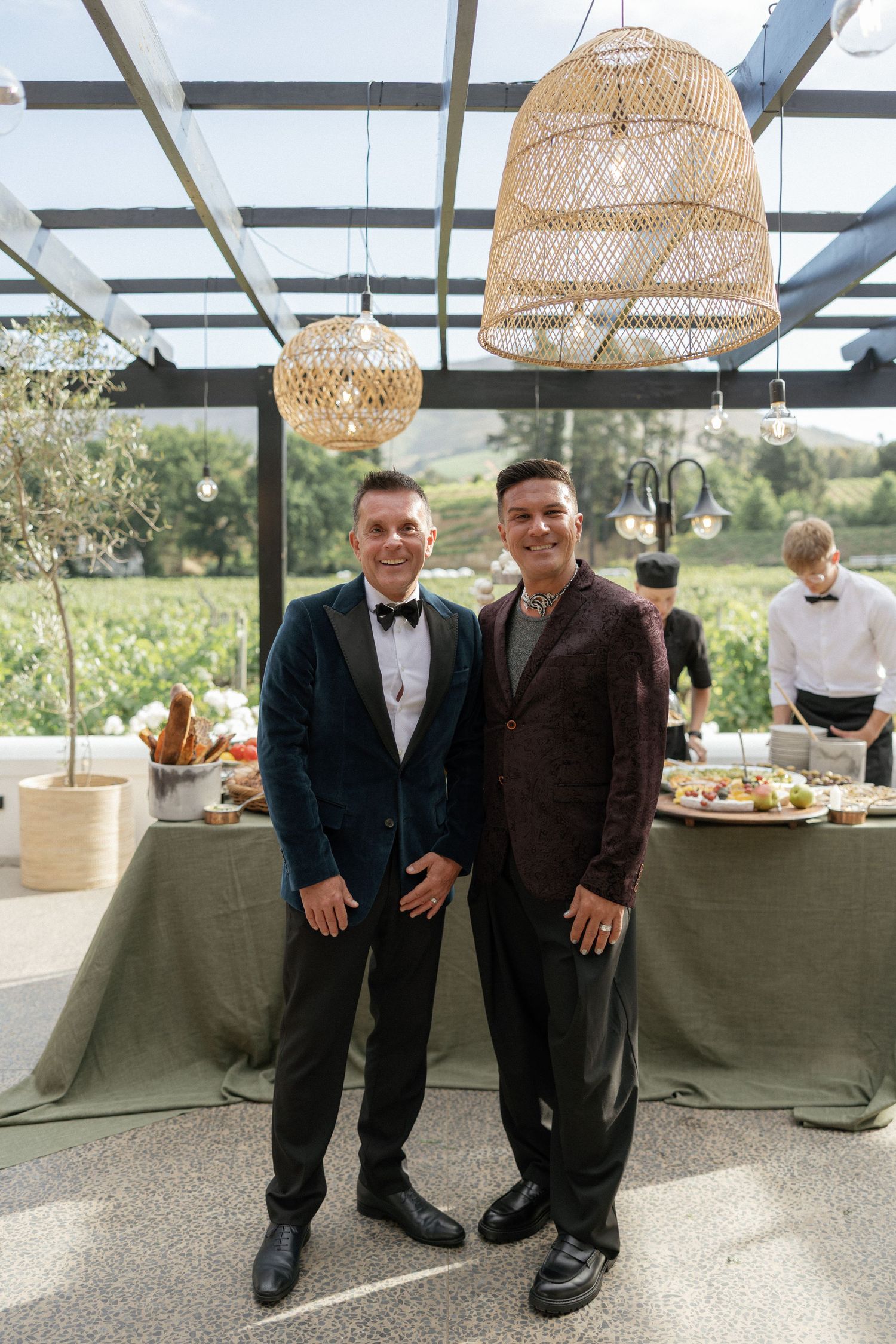 Two men in elegant blazers smile at a wedding reception with wicker pendant lights and a buffet table in the background.