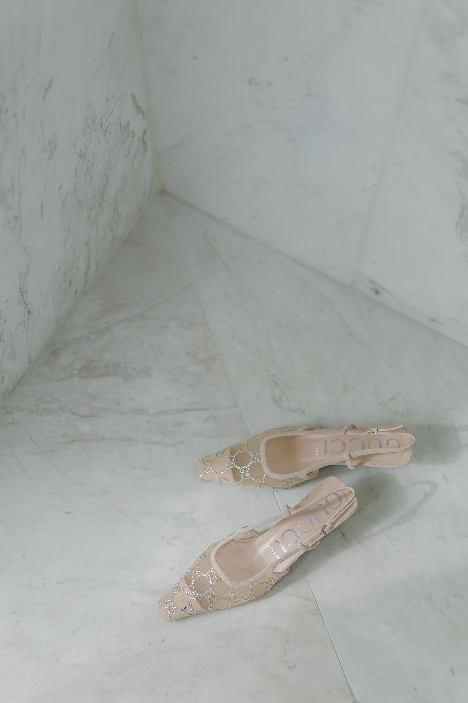 Pair of beige Gucci lace slingback heels placed on white marble floor in minimalist corner setting.