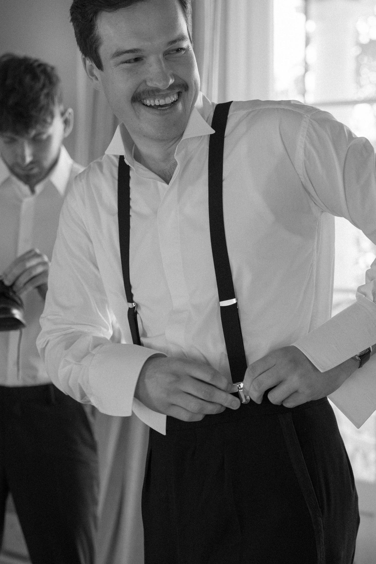 Smiling groom in white shirt and black suspenders getting ready in black and white photo.