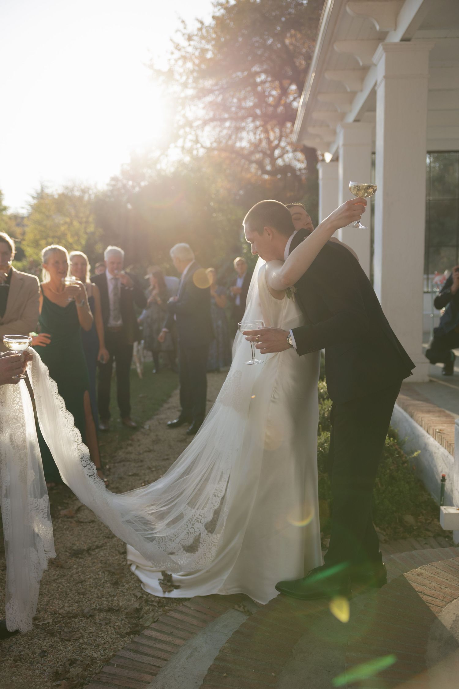 Bride and groom embracing and raising champagne glasses at golden hour outdoor wedding reception.