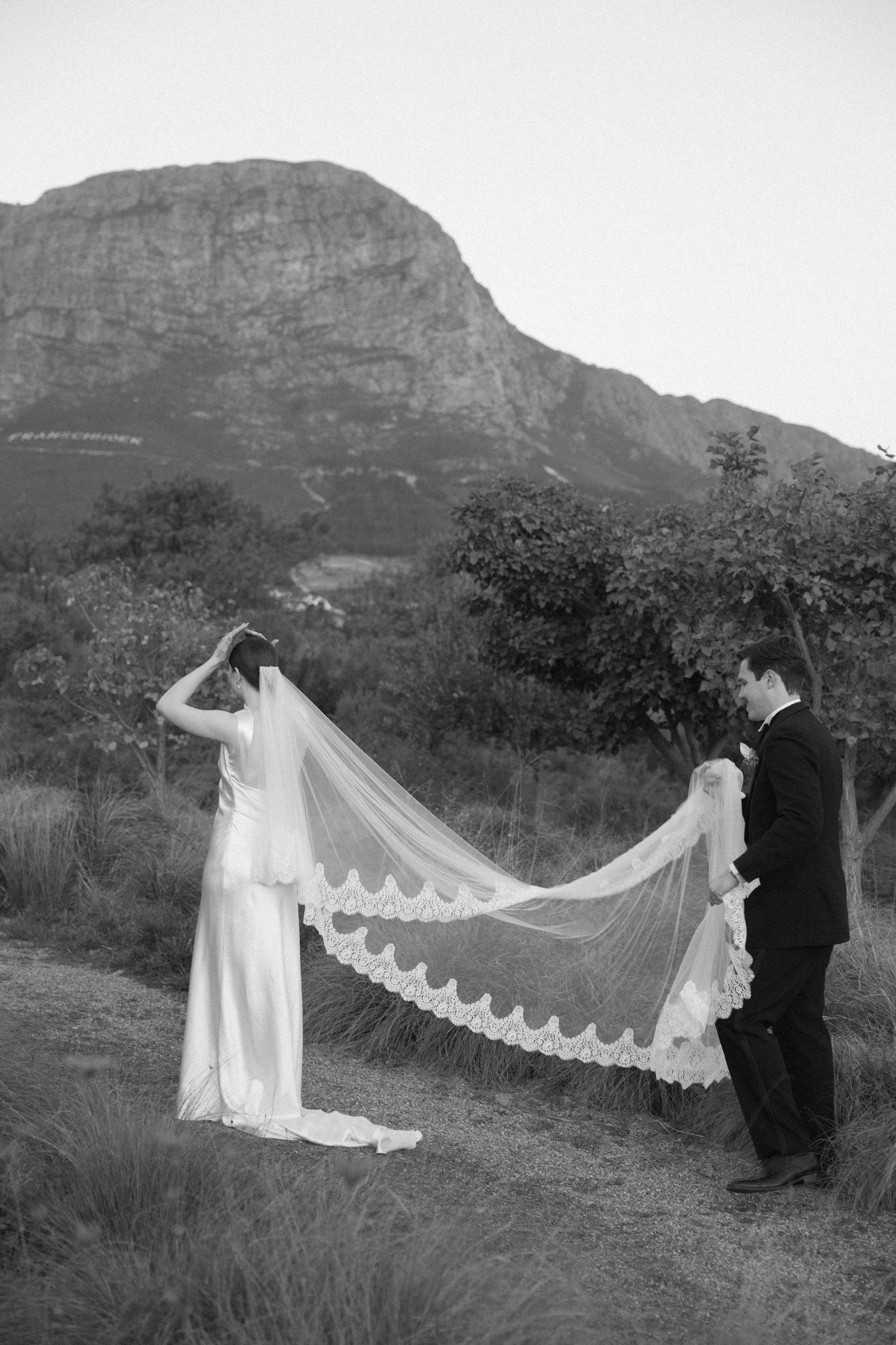 Black and white photo of groom holding bride's flowing lace-edged cathedral veil with mountain scenery.