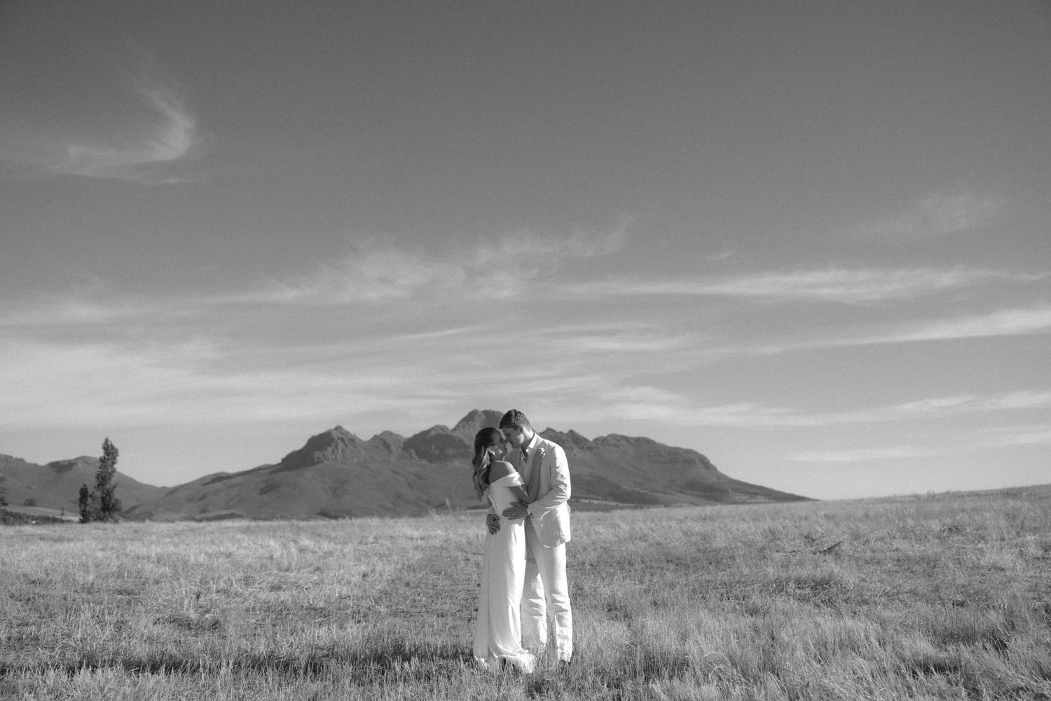 Black and white photo of couple embracing in open field with mountain backdrop.