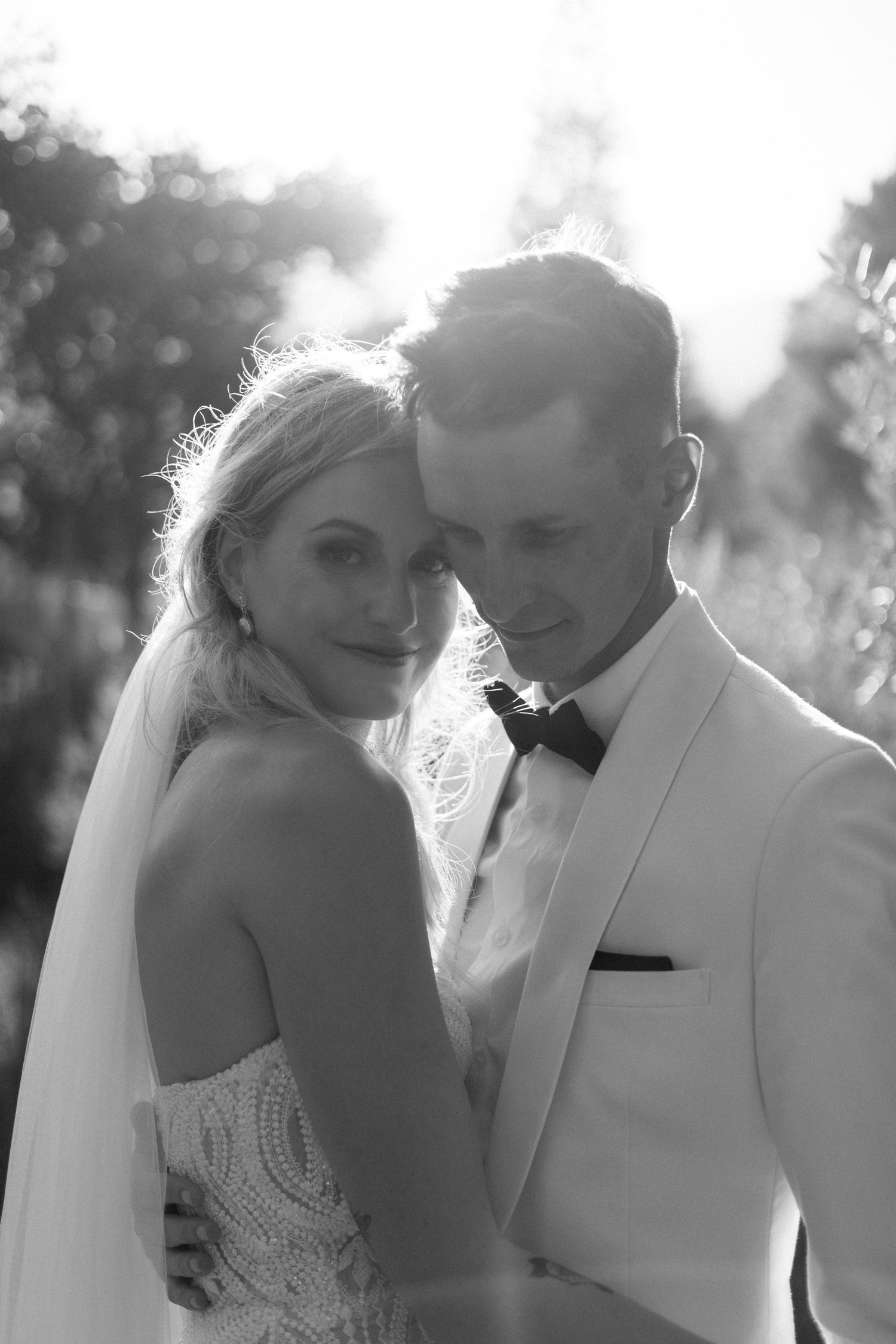 Black and white portrait of smiling bride leaning on groom in white tuxedo outdoors.