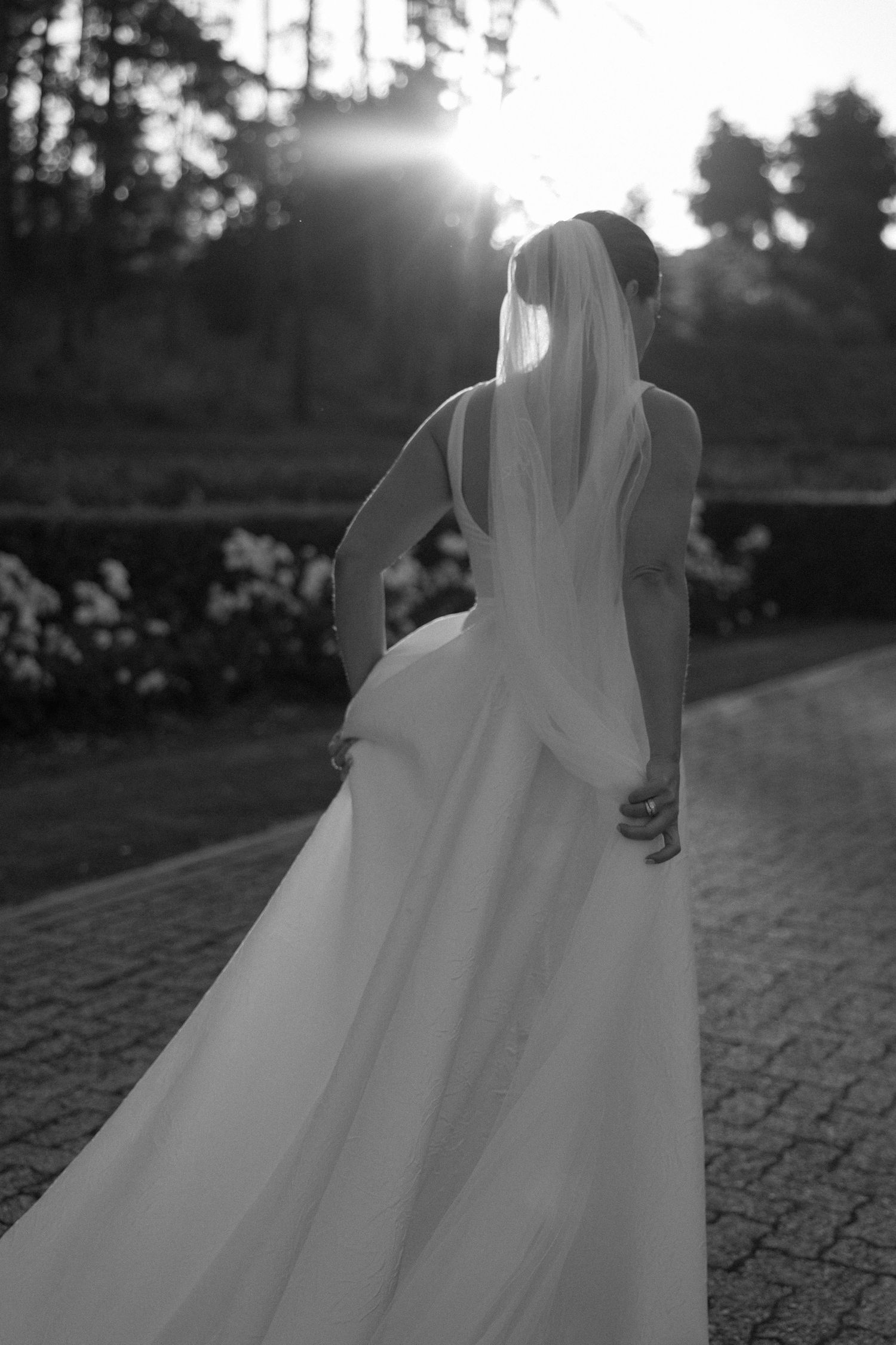 Black and white photo of a bride in a flowing wedding gown and veil, walking away toward a sunlit garden.