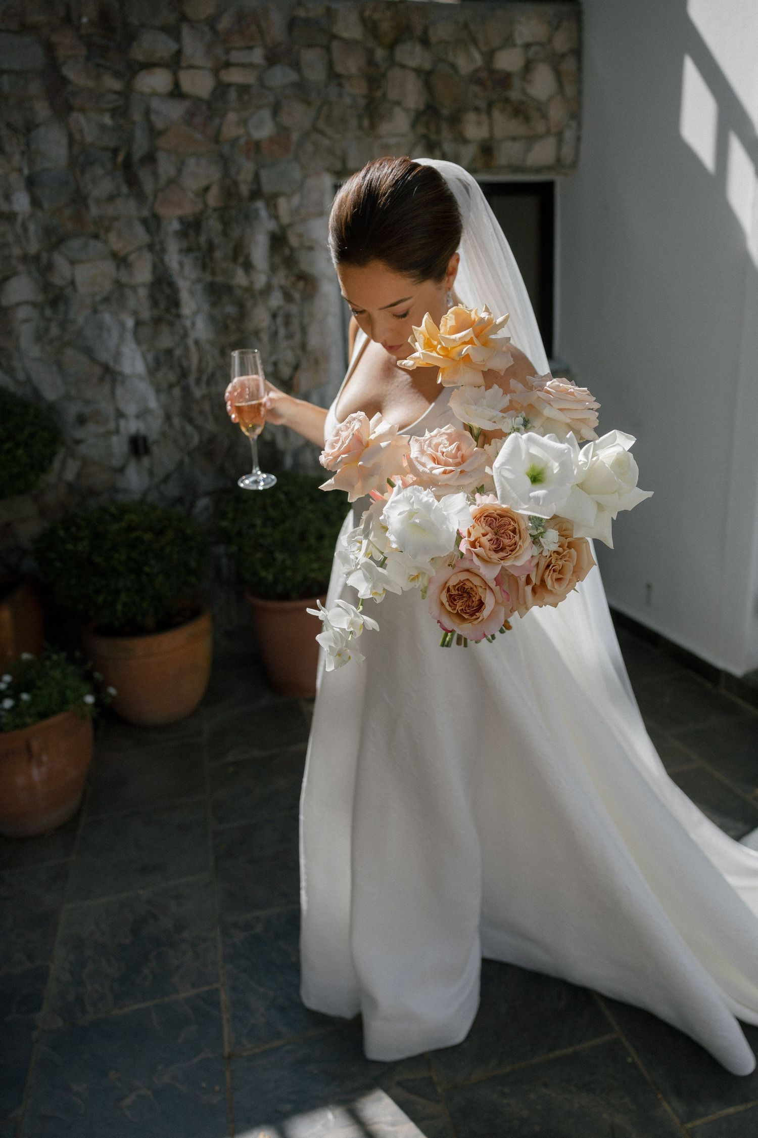Bride in white gown holds champagne glass and large peach and white floral bouquet outdoors near stone wall.