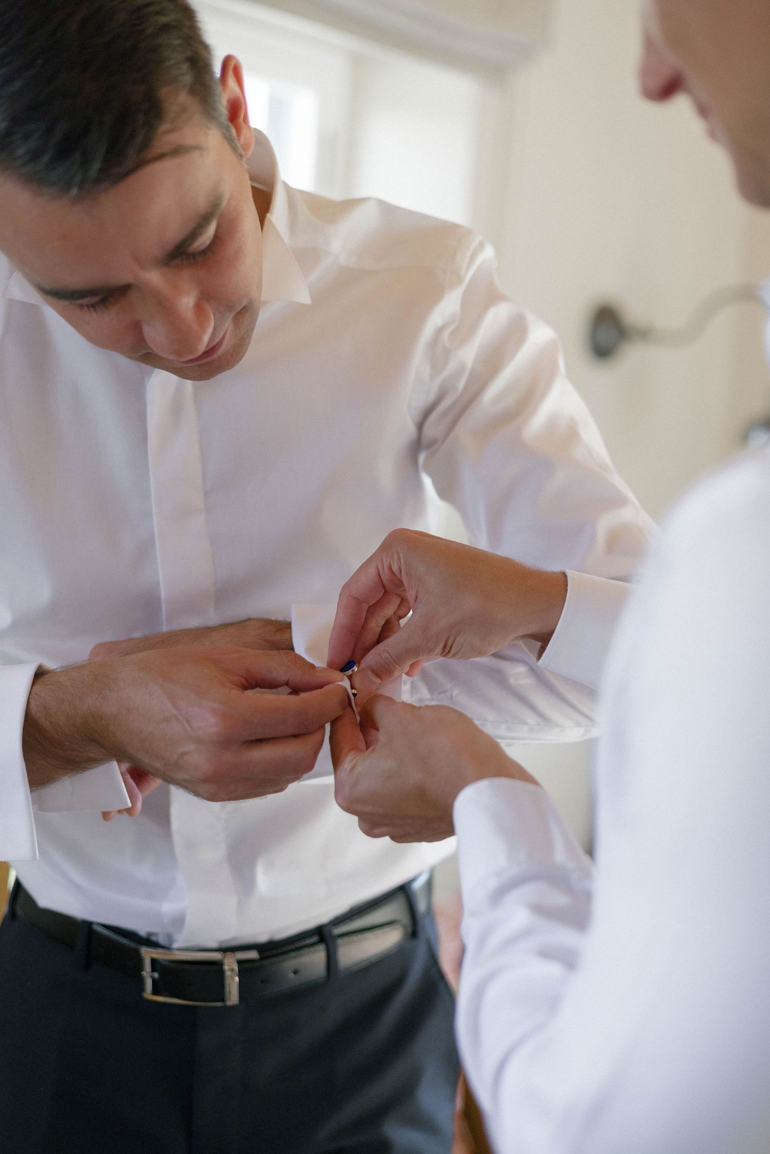 Groom being helped with cufflinks by groomsman while getting dressed for wedding ceremony.