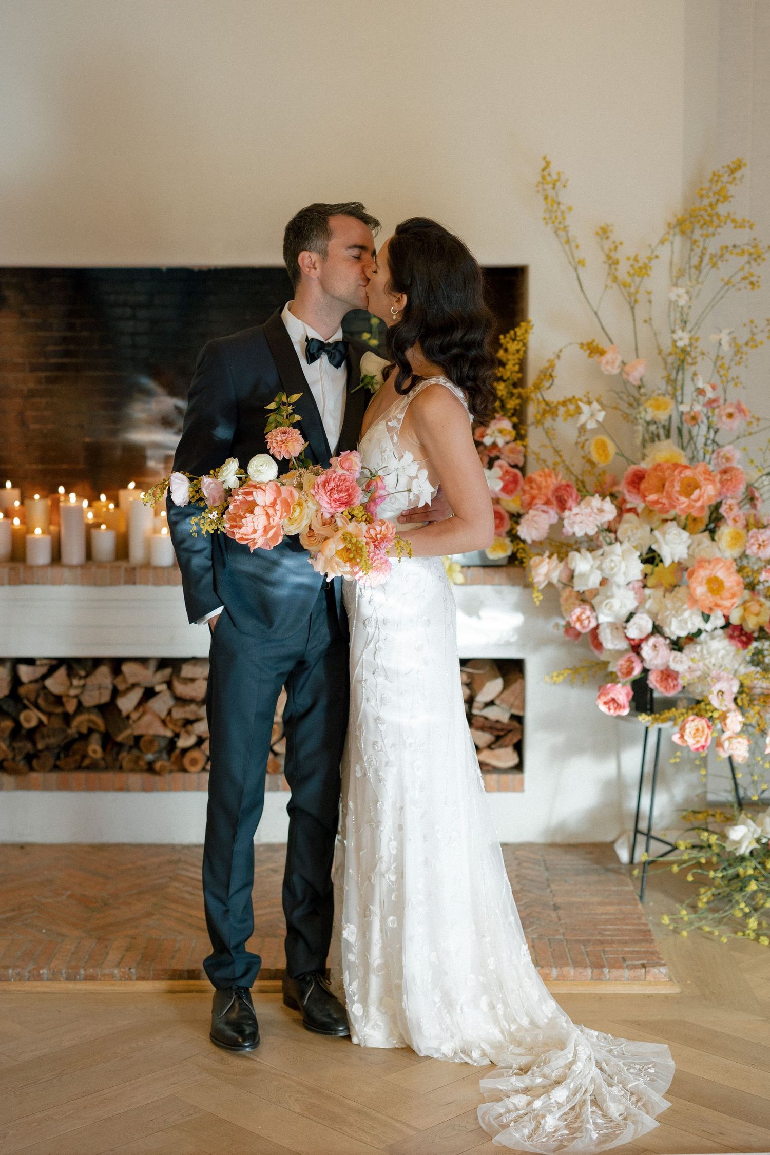 Bride and groom kissing near fireplace with candles and colorful floral arrangements.