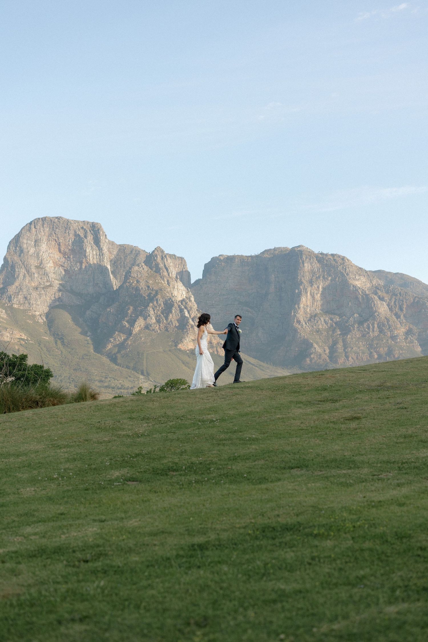 Bride and groom walking hand in hand up green hillside with majestic rocky mountains in background.