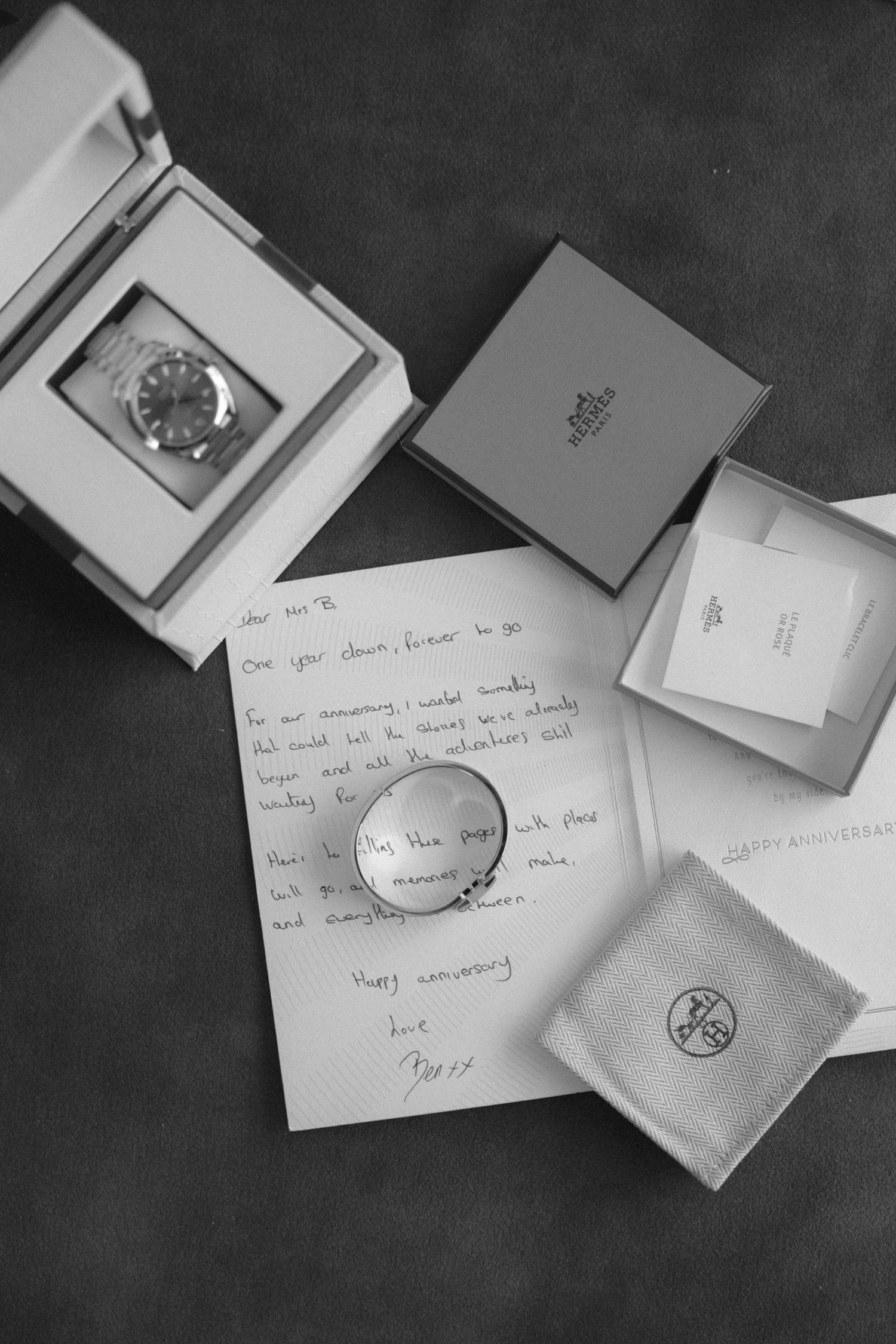 Black and white flat lay of anniversary gifts including a luxury watch, Hermes boxes, a ring, and handwritten love letter.