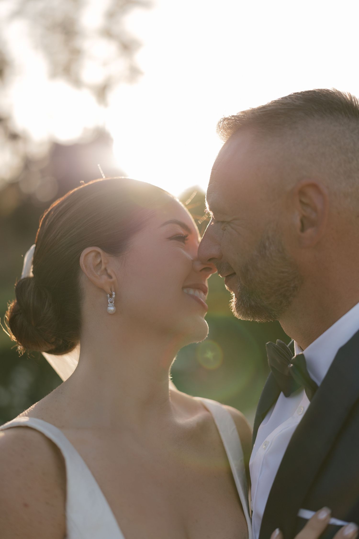 A bride and groom share a romantic nose-to-nose moment bathed in golden sunset light on their wedding day.