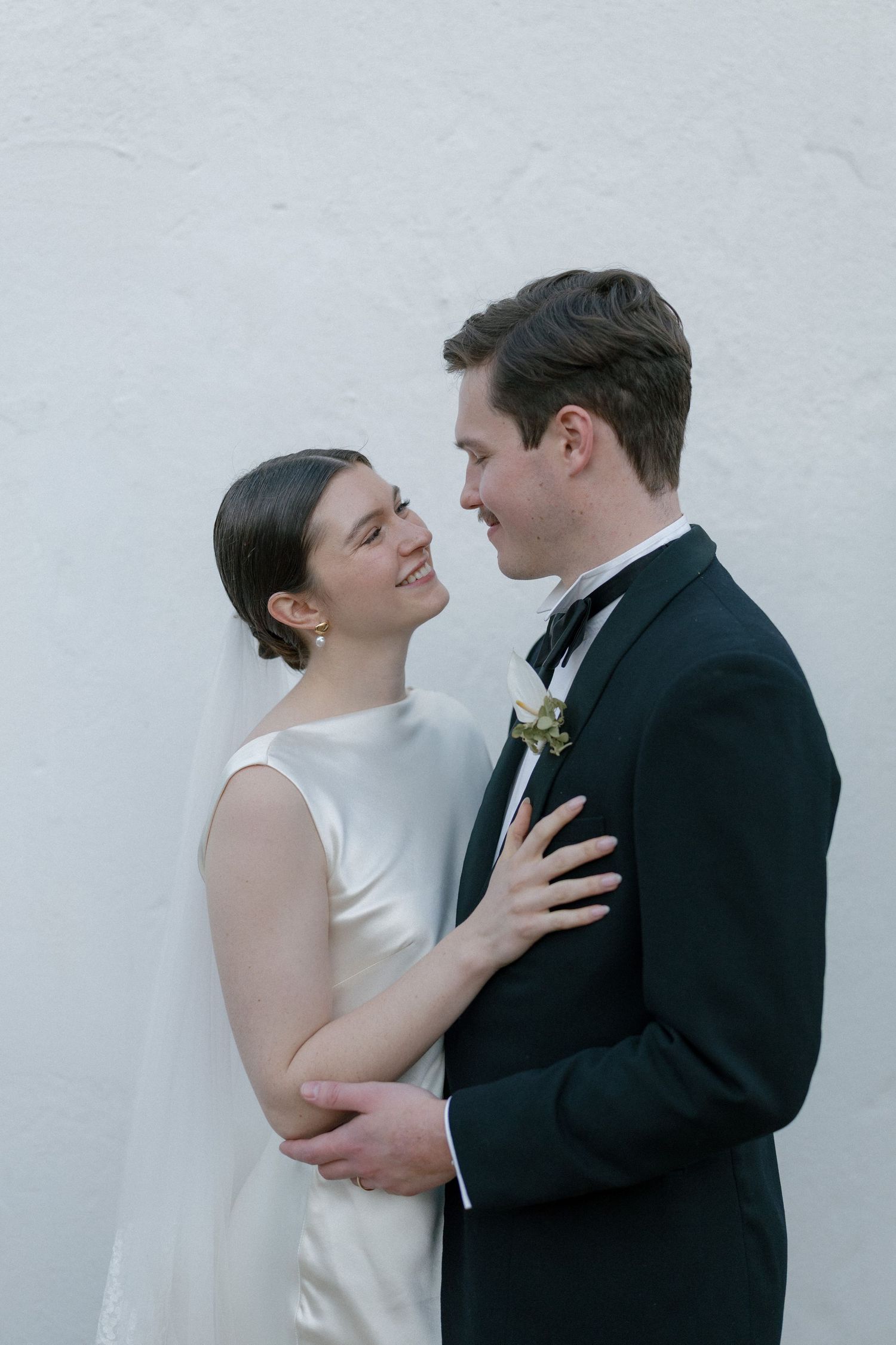 Bride and groom sharing an intimate moment against a white wall, dressed in classic wedding attire.