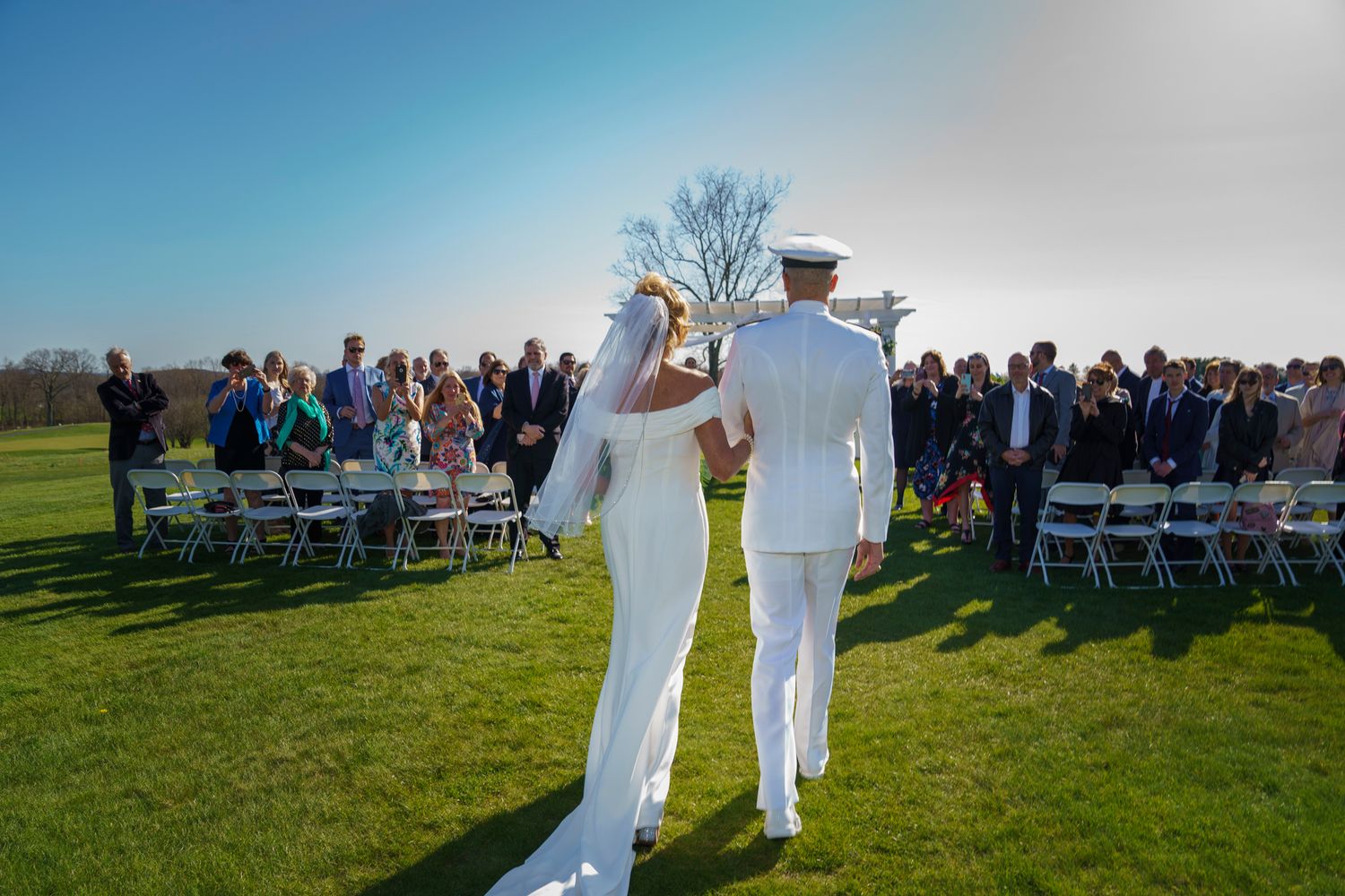 Military groom in white uniform walks bride down outdoor wedding aisle on sunny green lawn with seated guests.
