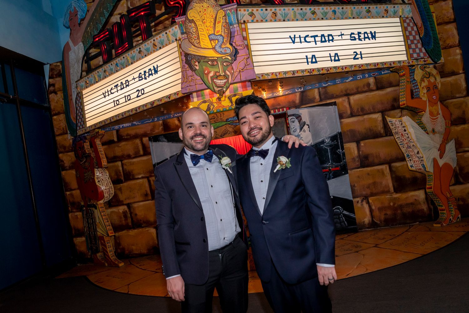 Two grooms pose smiling in front of a theater marquee displaying their names Victor and Sean with wedding date 10-14-21.
