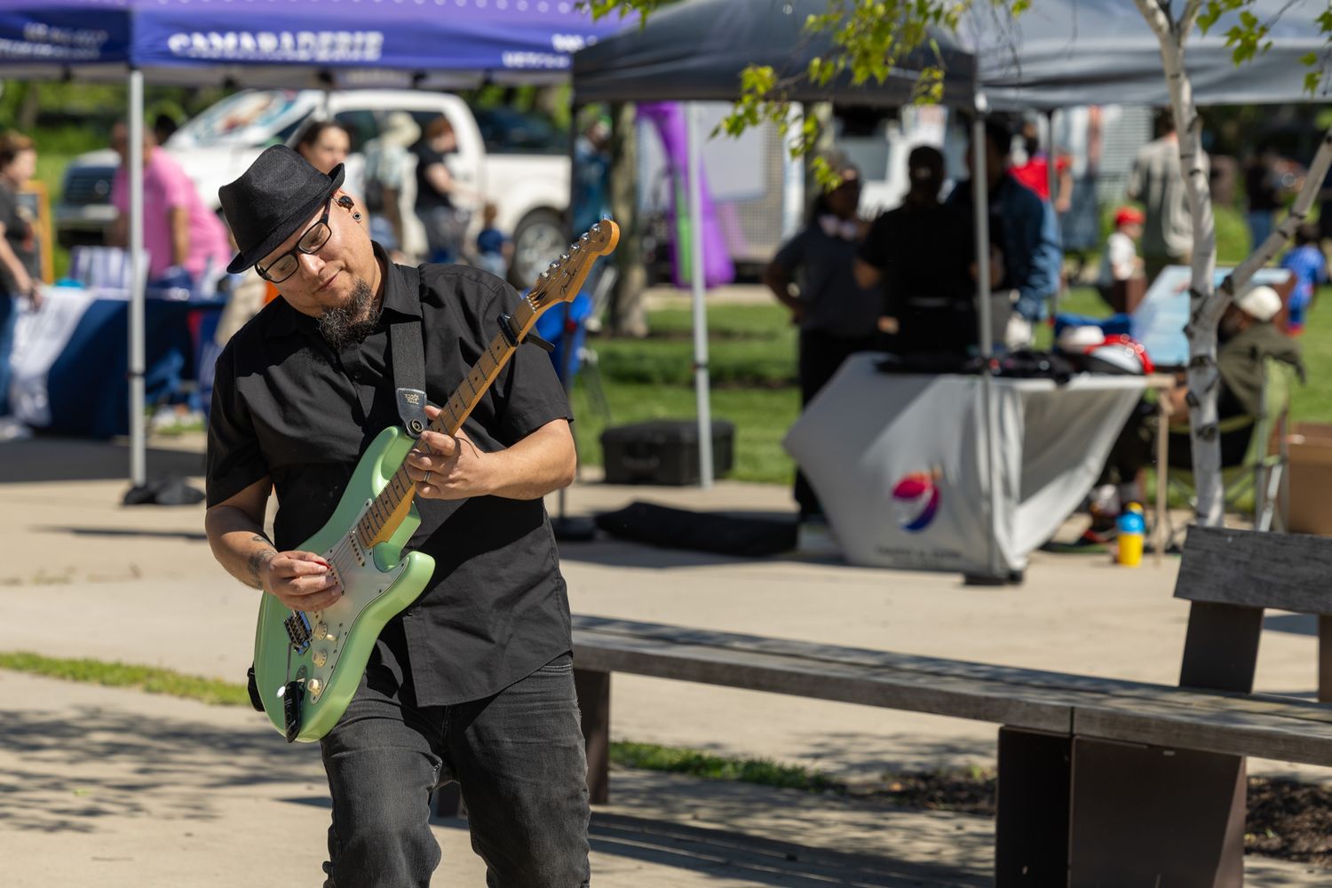 Man in black hat plays green electric guitar outdoors at a sunny community event with vendor tents in background.