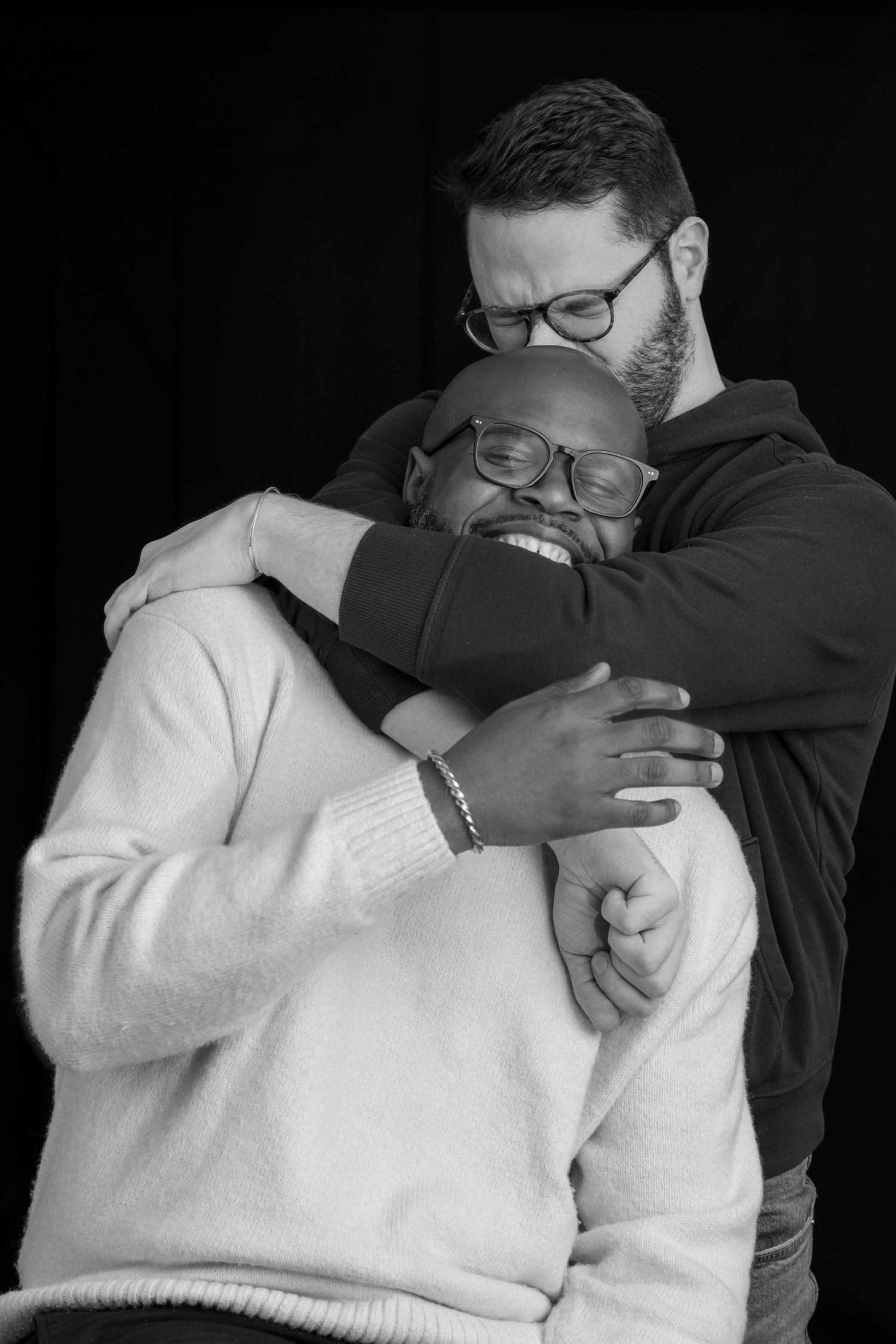 Two men sharing a warm embrace, smiling, in a black and white studio portrait.