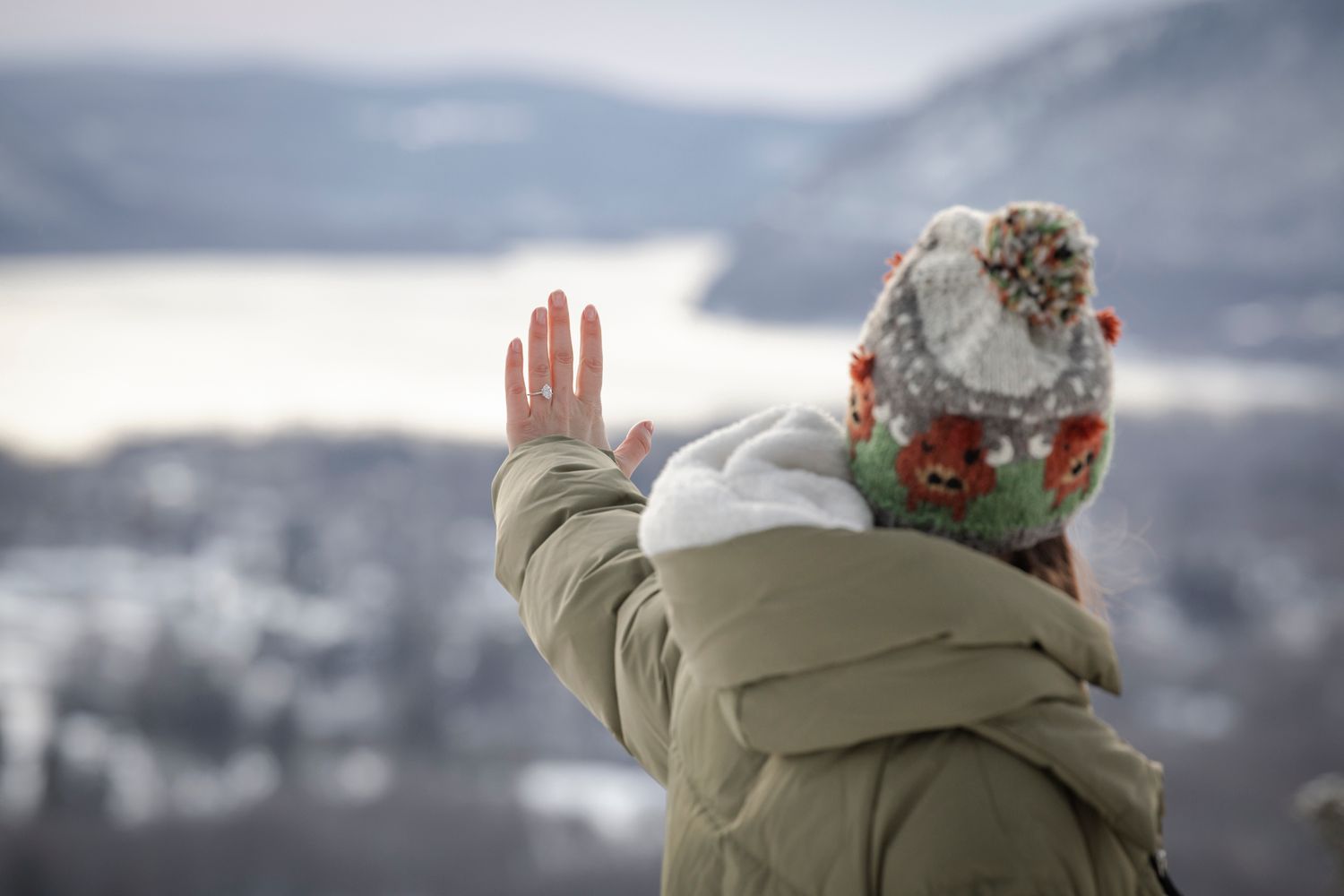 Person in winter jacket and colorful knit hat waving hand with snowy mountain landscape in background.