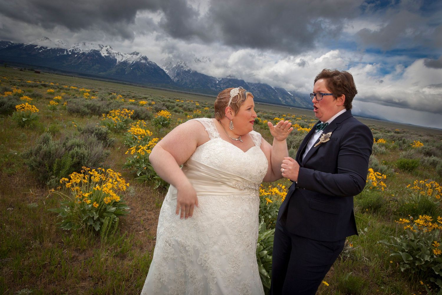 Two brides share a joyful moment in a wildflower meadow with snow-capped Teton mountains under dramatic cloudy skies.