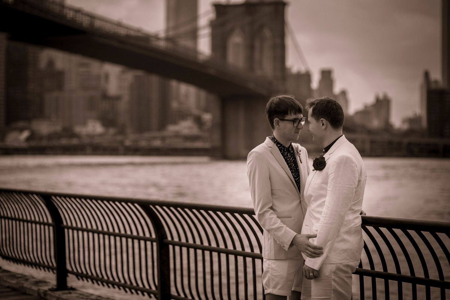 Two grooms in white suits hold hands by Brooklyn Bridge waterfront in a romantic sepia-toned wedding photo.