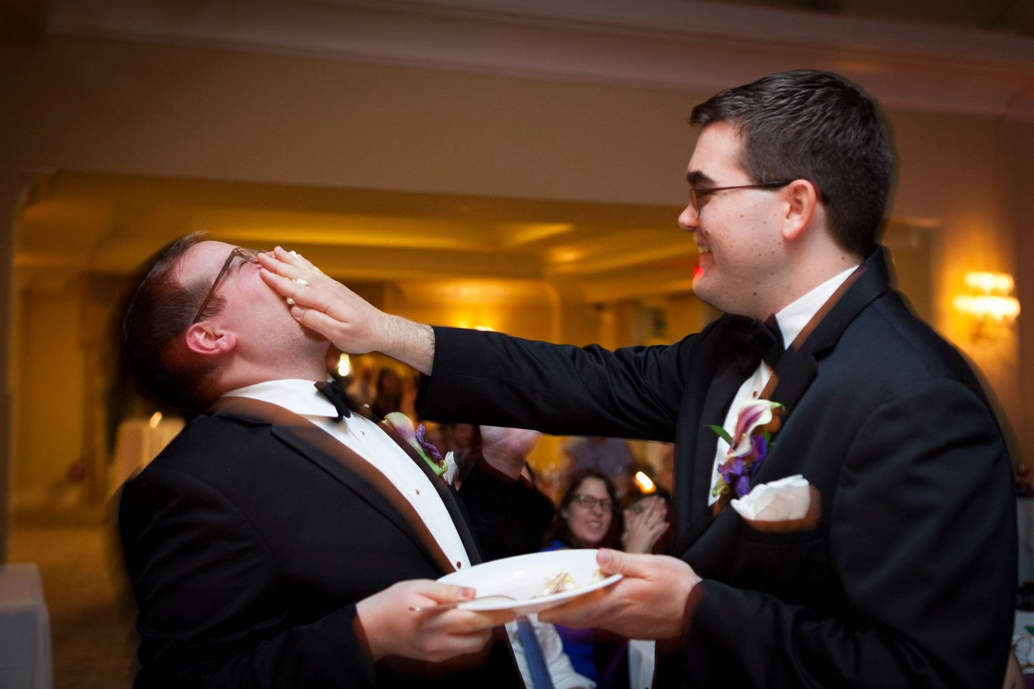 Two grooms in tuxedos sharing a fun cake cutting moment at their wedding reception.