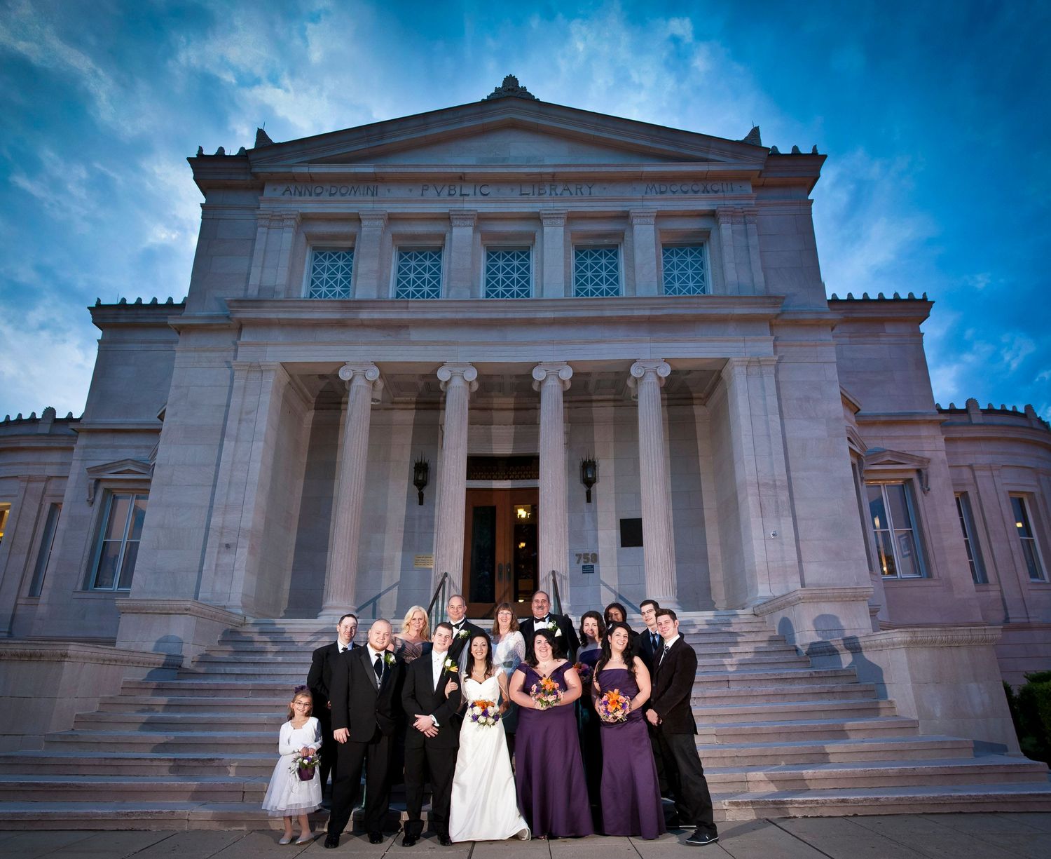A wedding party posing on the steps of a grand neoclassical building at dusk.