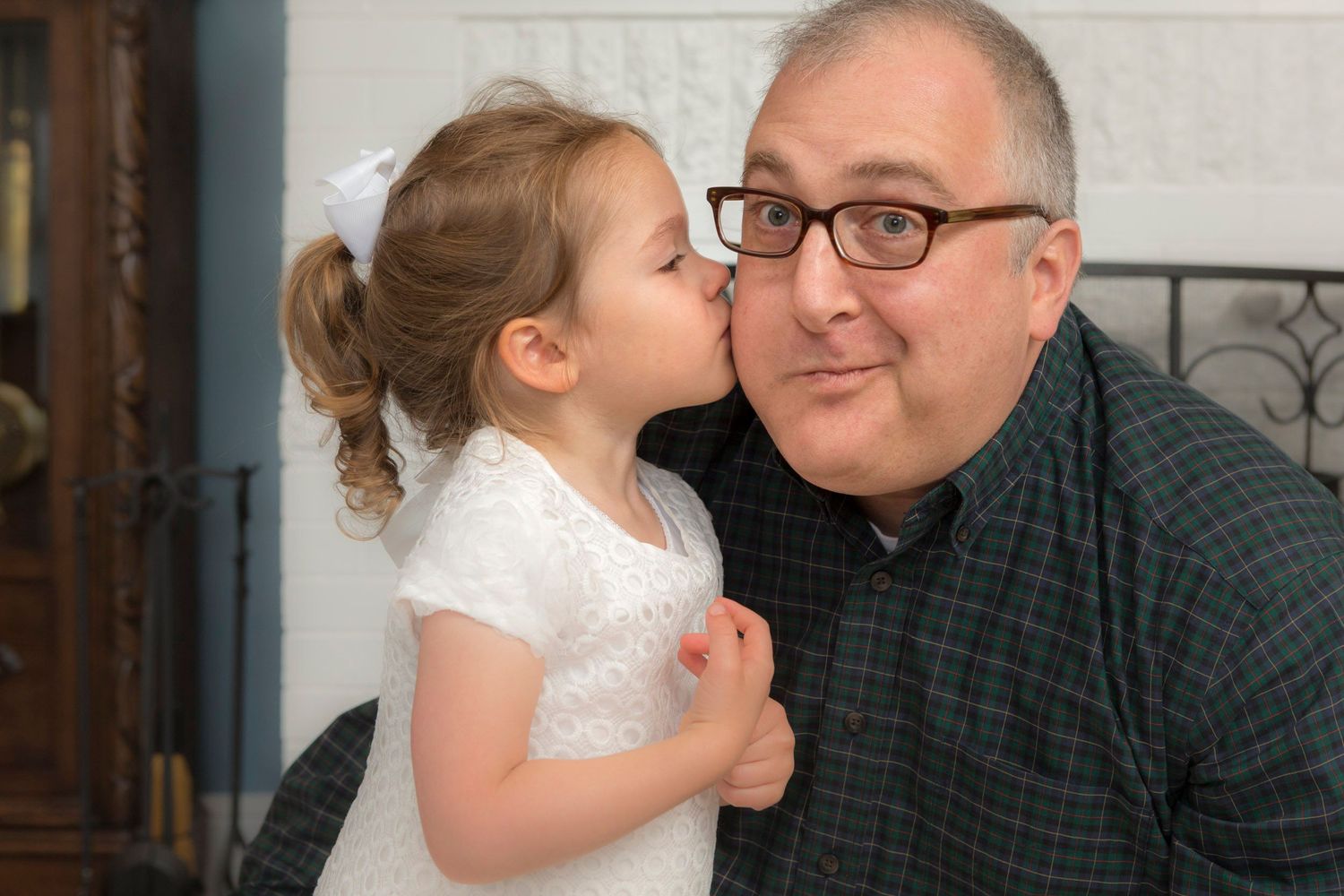 Young girl in white dress giving a kiss on the cheek to a smiling man with glasses.