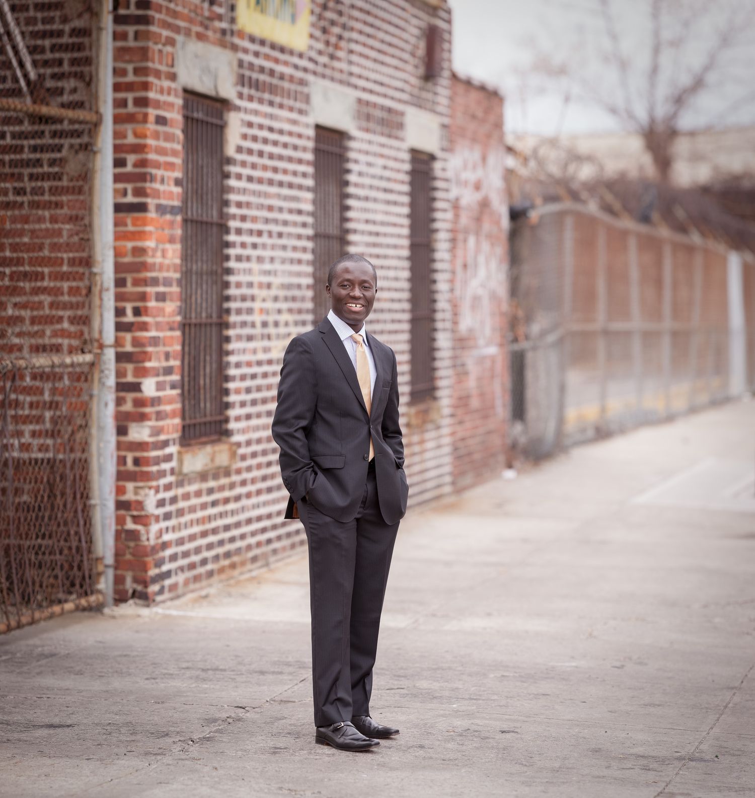 Smiling man in dark suit standing in urban alley beside brick building.