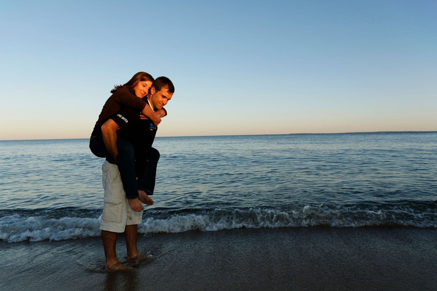 Groom giving bride a piggyback ride on beach at sunset by ocean waves.