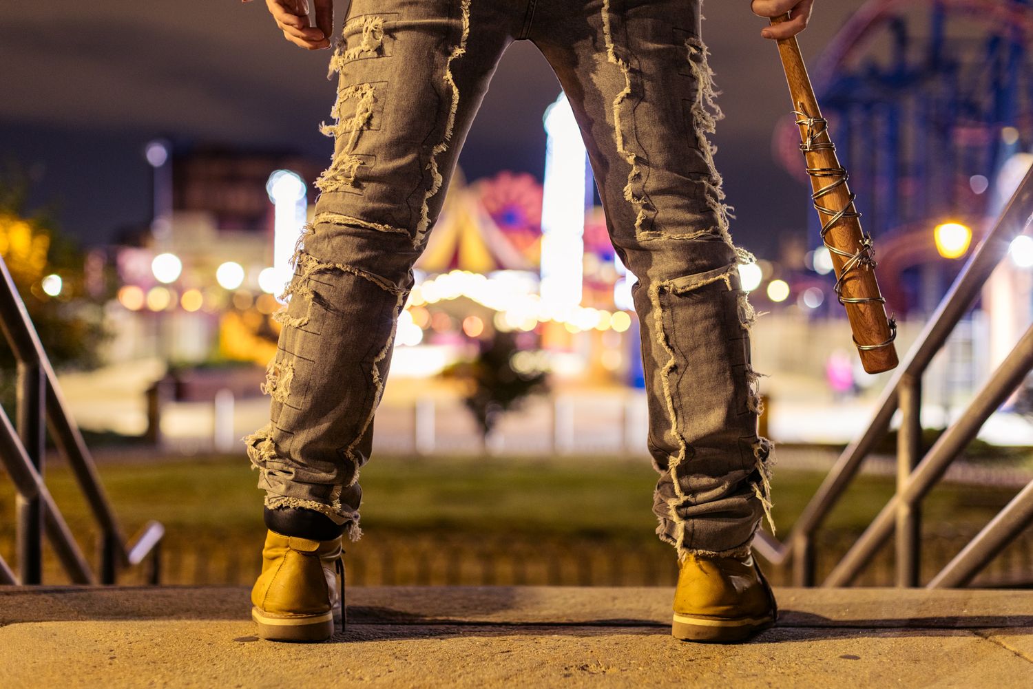 Person in ripped distressed jeans and tan work boots stands on a ledge holding a baseball bat near a glowing nighttime carnival.