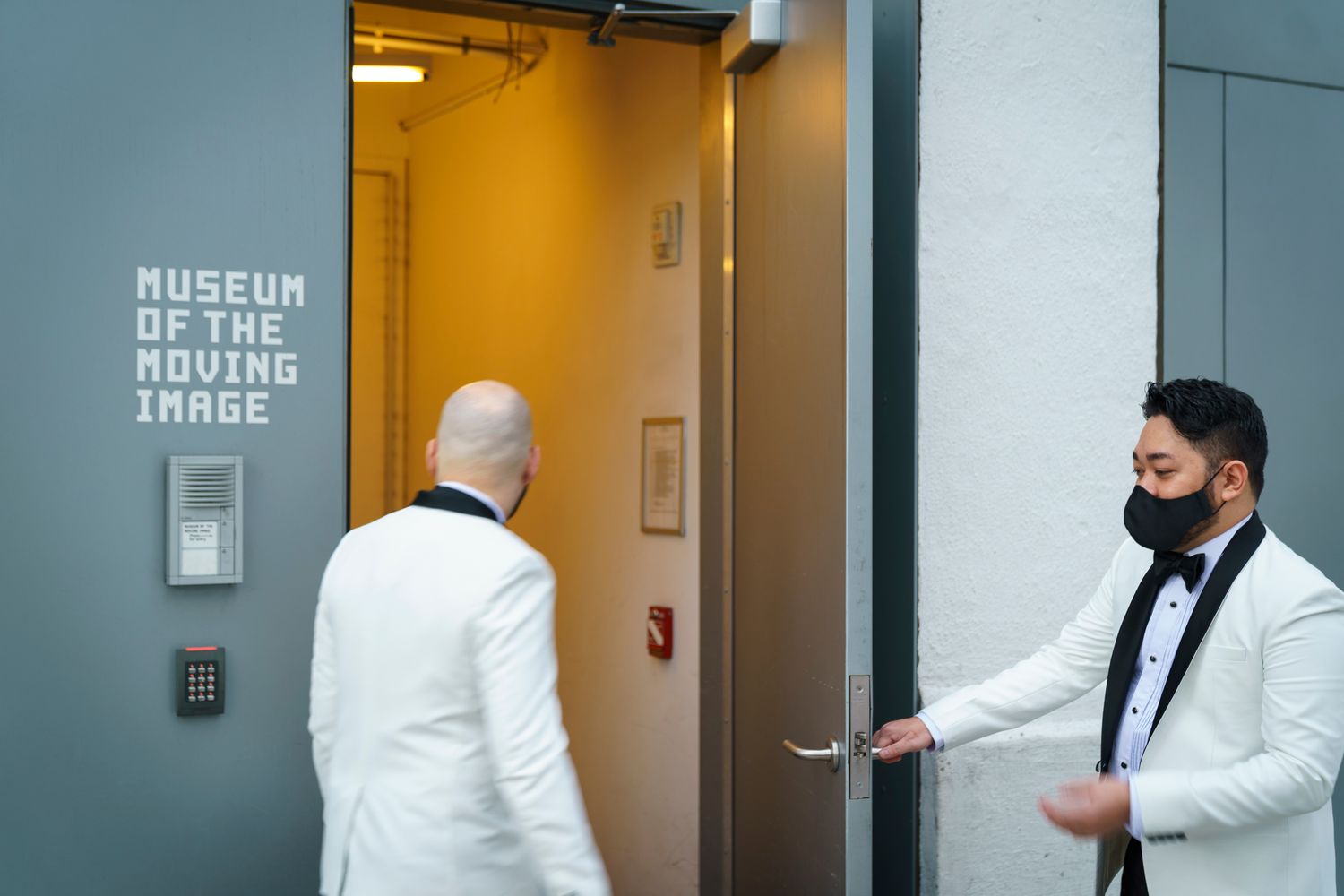 Two men in white tuxedos enter the Museum of the Moving Image, one holding the door open while wearing a black mask.