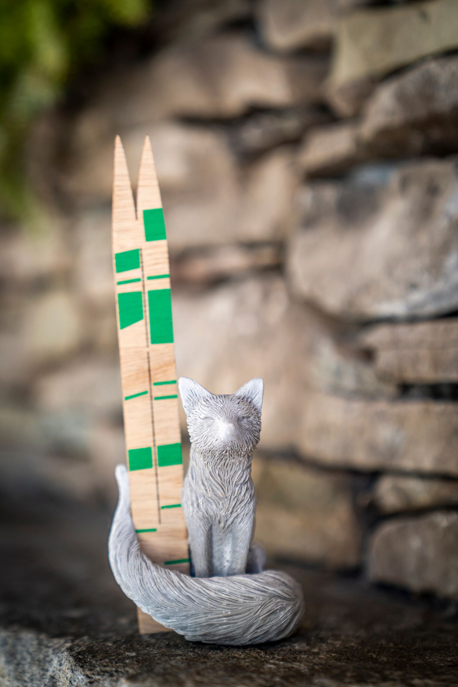 Gray fox figurine with wooden skis featuring green accents, posed on stone surface with blurred rock wall background.